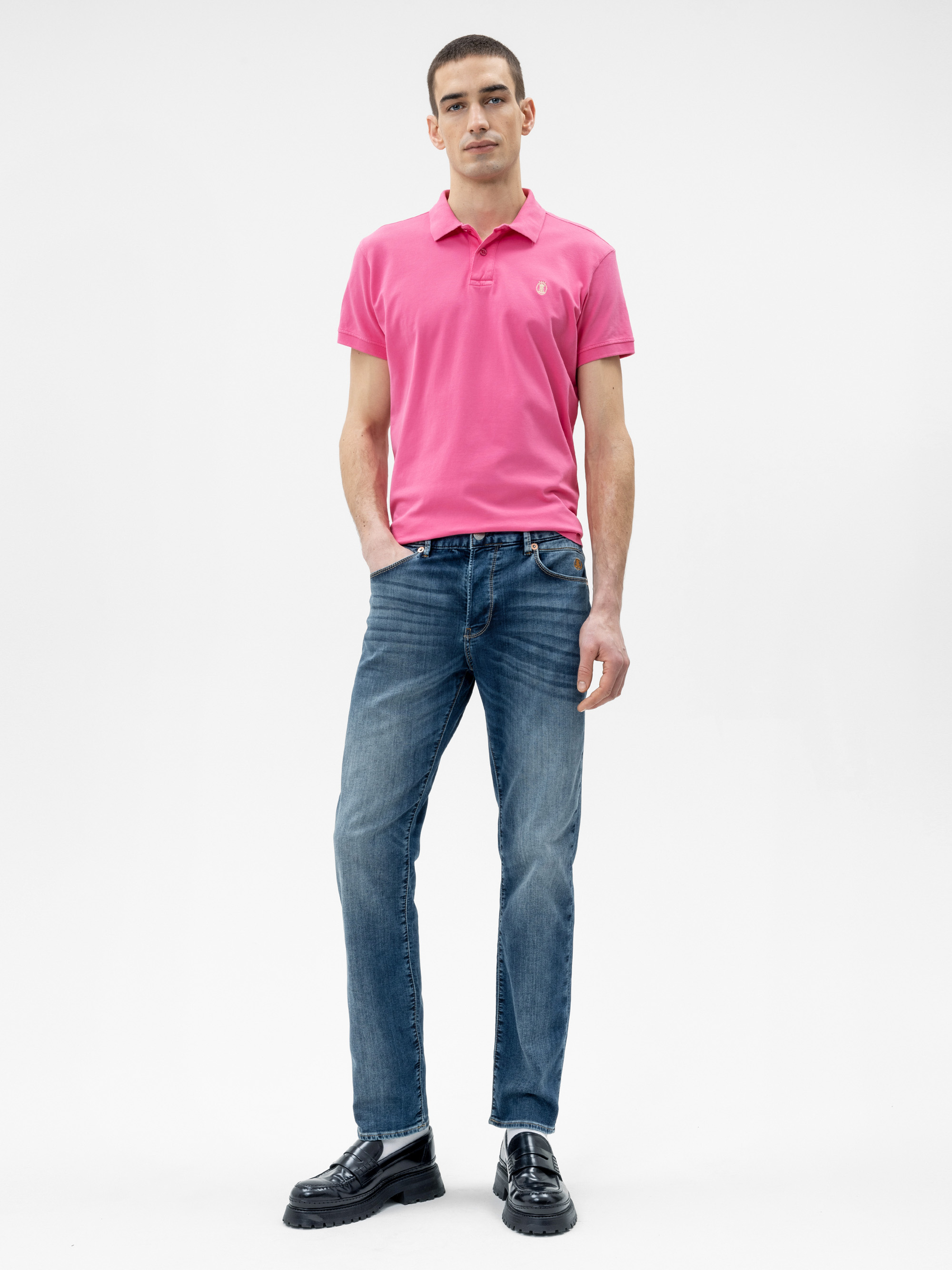 Ein Mann steht vor einem schlichten weißen Hintergrund, trägt ein hellrosa Polohemd, blaue Jeans und schwarze Slipper, eine Hand in der Tasche und einen neutralen Gesichtsausdruck.