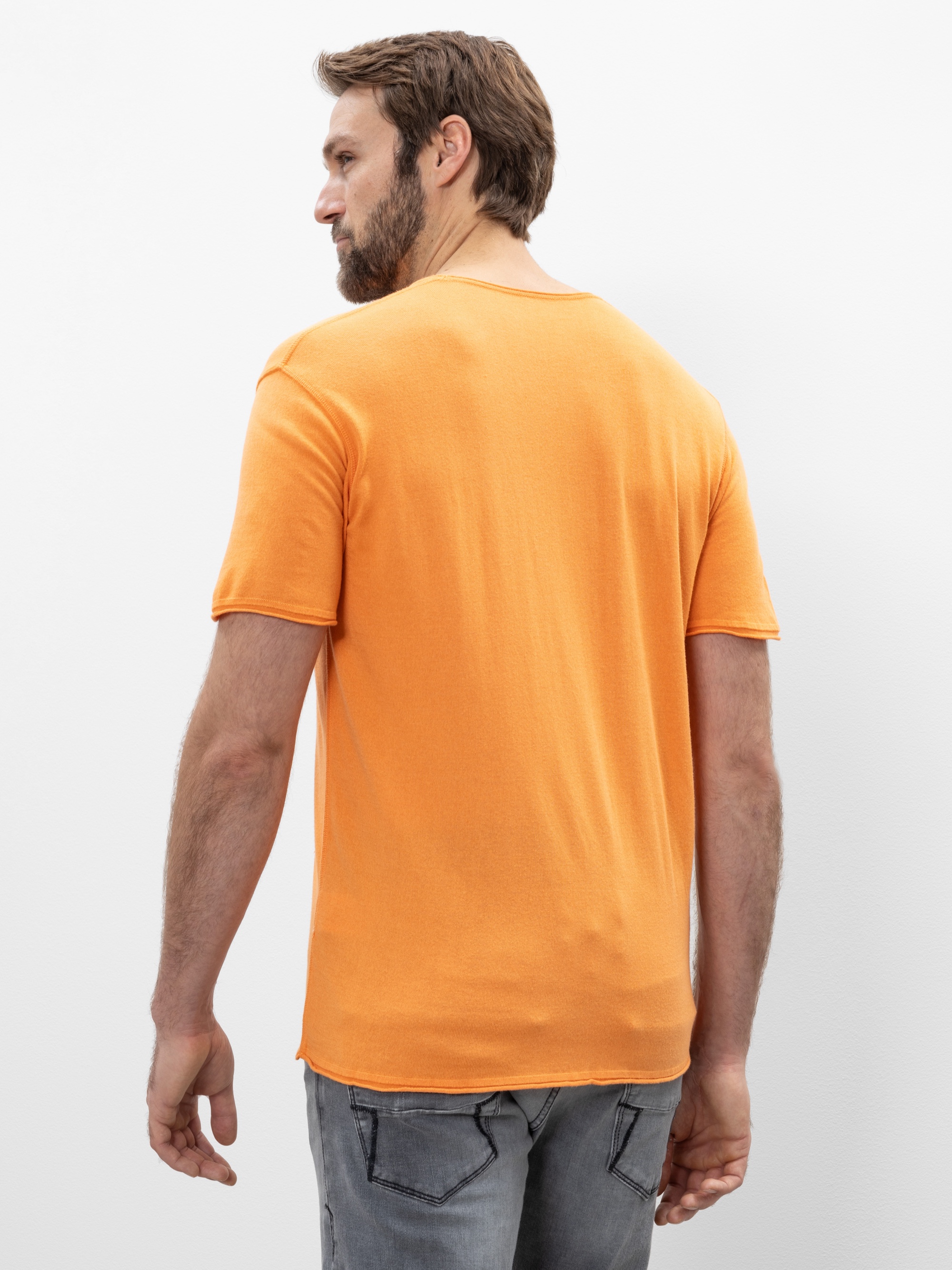 Ein Mann mit braunen Haaren und Bart steht mit dem Gesicht zur Wand, trägt ein orangefarbenes, kurzärmeliges T-Shirt und hellgraue Jeans vor einem schlichten weißen Hintergrund.