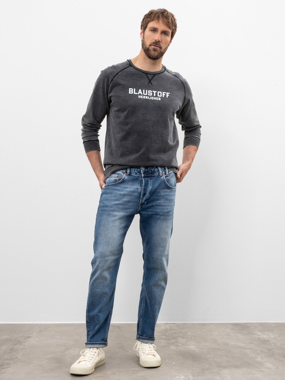 Ein Mann mit Bart trägt ein kohlegraues BLAUSTOFF-Sweatshirt, hellblaue Jeans und weiße Turnschuhe. Er steht in einem Innenraum vor einer schlichten weißen Wand und schaut mit den Händen in den Taschen in die Kamera.