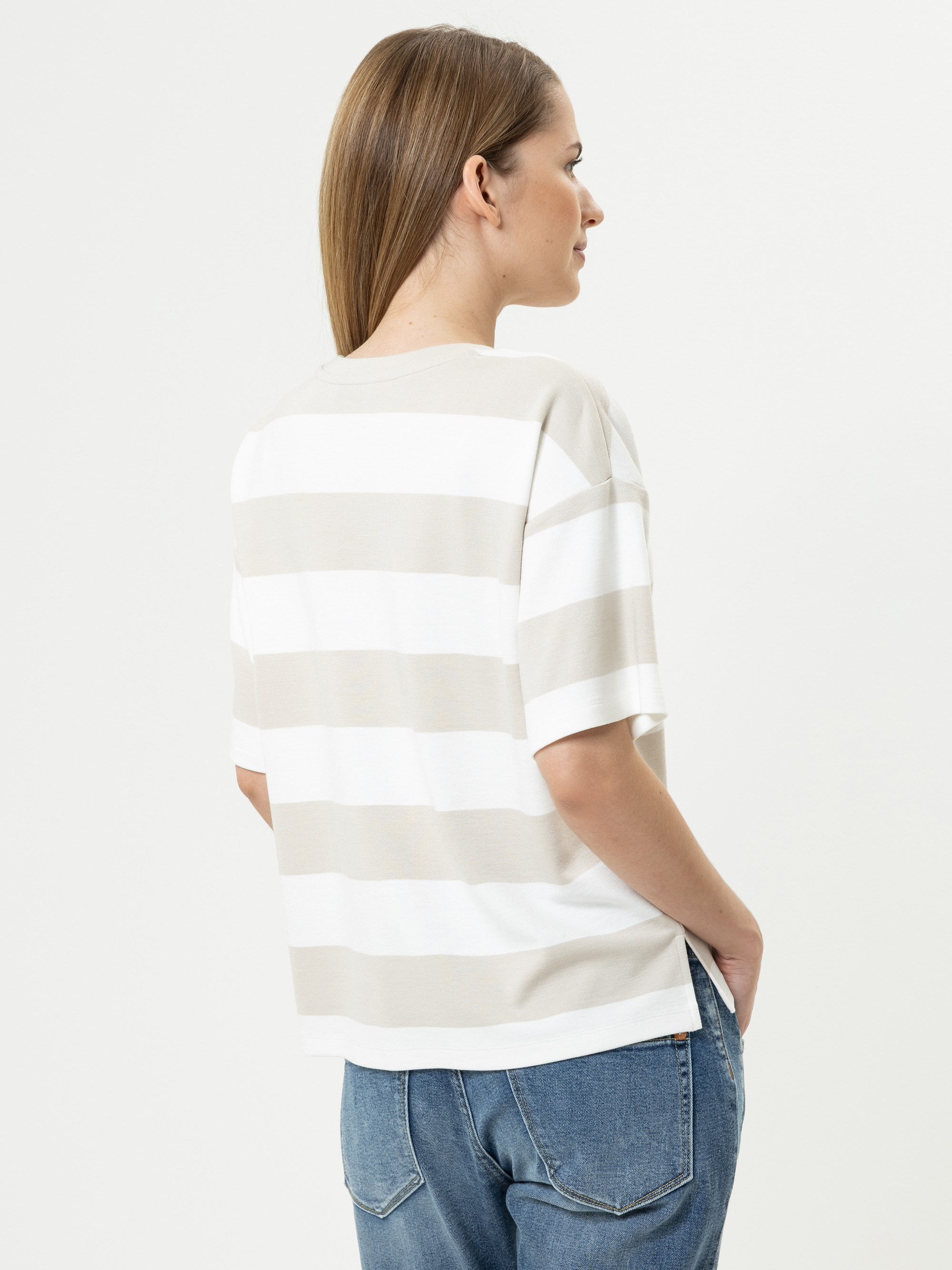 Eine Frau mit glattem, hellbraunem Haar trägt ein lockeres, kurzärmeliges, beige-weiß gestreiftes Hemd und blaue Jeans. Sie steht mit den Händen in den Taschen, leicht von der Kamera abgewandt.