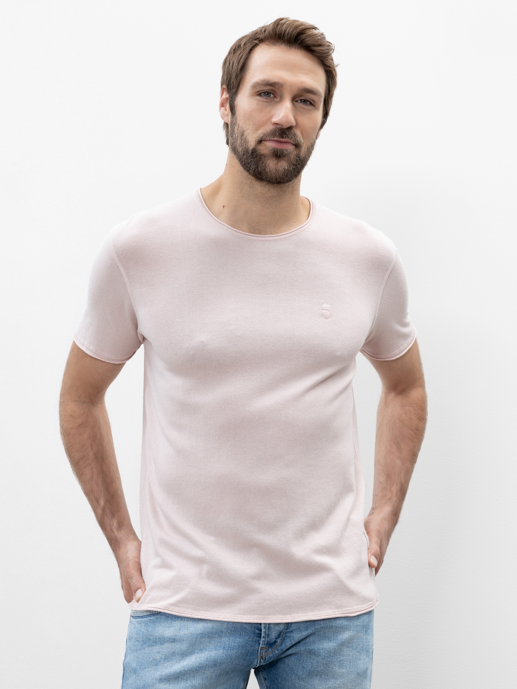 Vor einem schlichten weißen Hintergrund steht ein Mann mit kurzen braunen Haaren und Bart, der ein hellrosa kurzärmeliges T-Shirt und hellblaue Jeans trägt und die Hände in den Taschen hat.