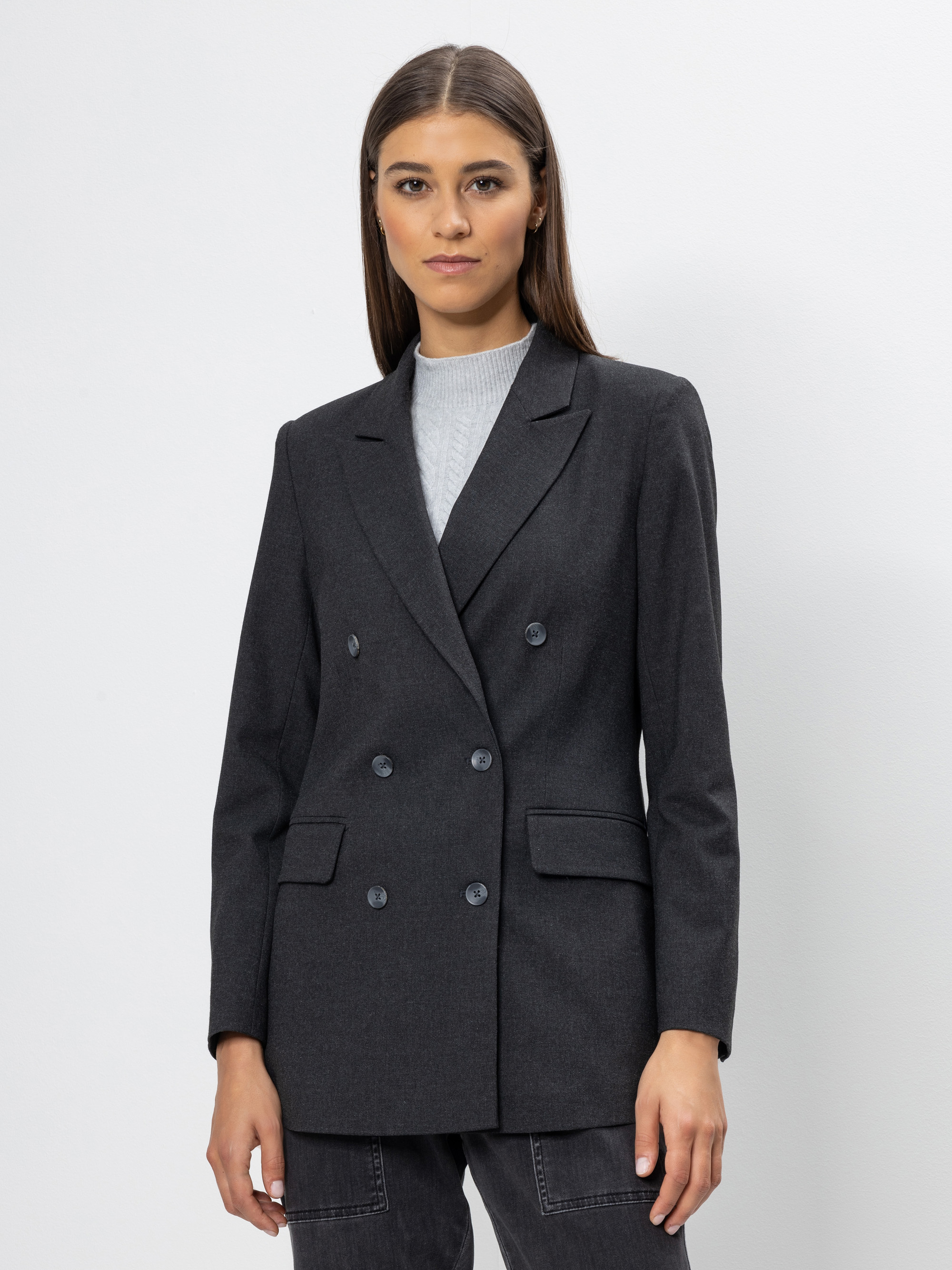 Eine Frau mit glattem braunem Haar trägt einen dunkelgrauen zweireihigen Blazer über einem hellgrauen Oberteil, steht vor einem schlichten weißen Hintergrund und blickt direkt in die Kamera.