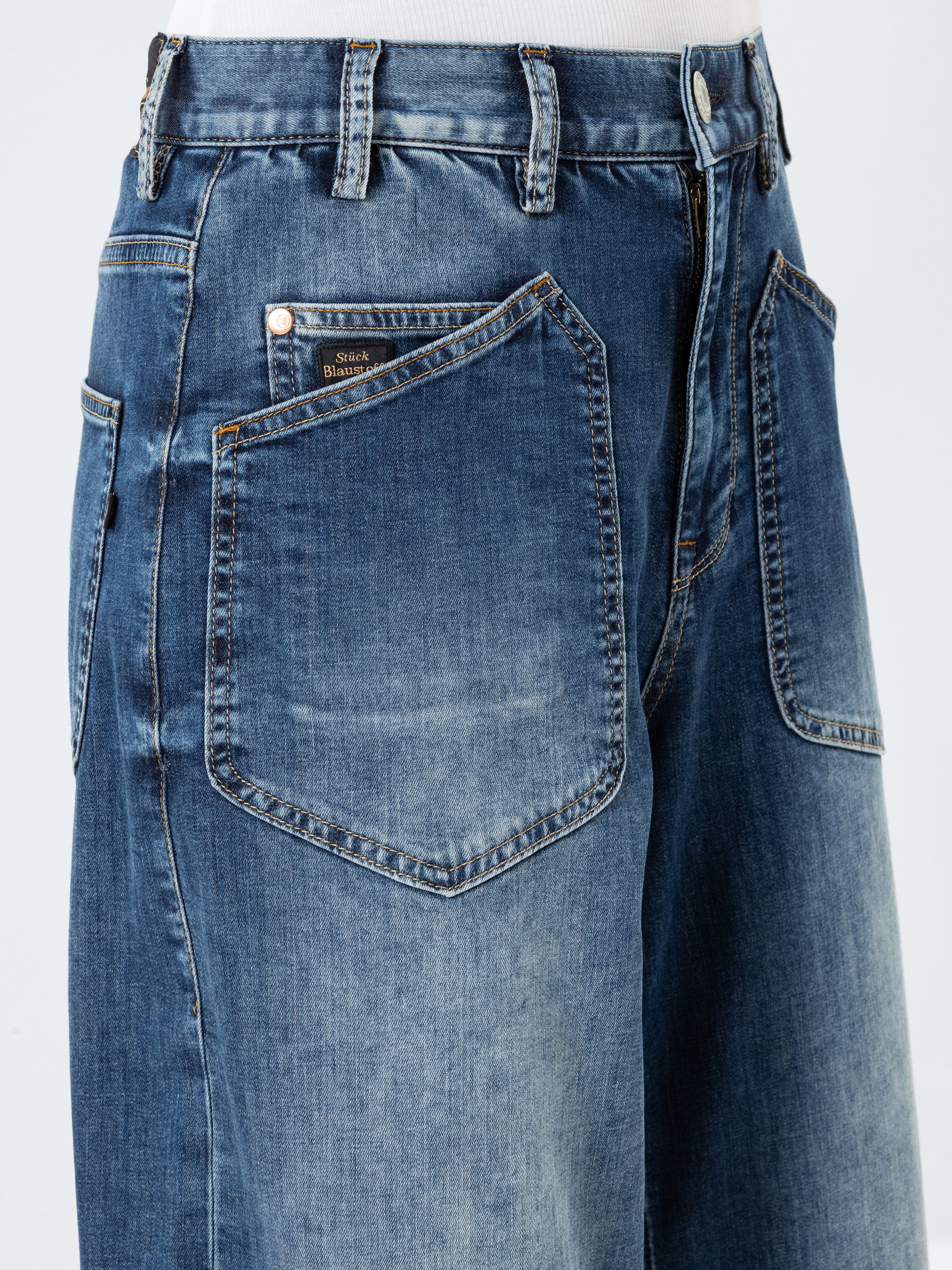Nahaufnahme einer blauen Denim-Jeans mit großen aufgesetzten Taschen vorne, einer Münztasche mit Etikett, orangefarbenen Nähten und einem teilweise sichtbaren Reißverschluss, getragen mit einem weißen Oberteil.