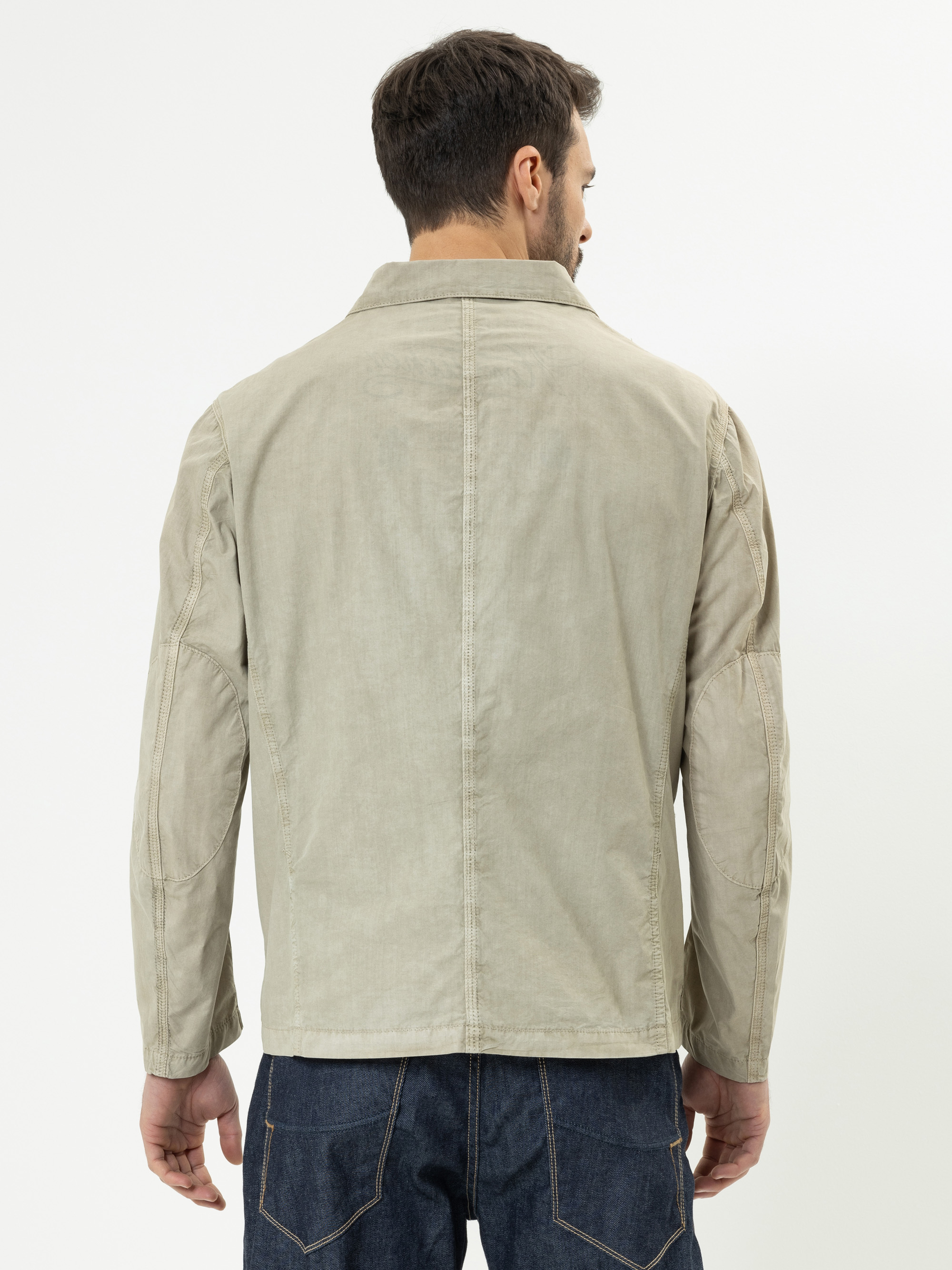 Ein Mann steht mit dem Rücken zur Kamera, trägt eine hellbeige Jacke und dunkelblaue Jeans vor einem schlichten weißen Hintergrund.