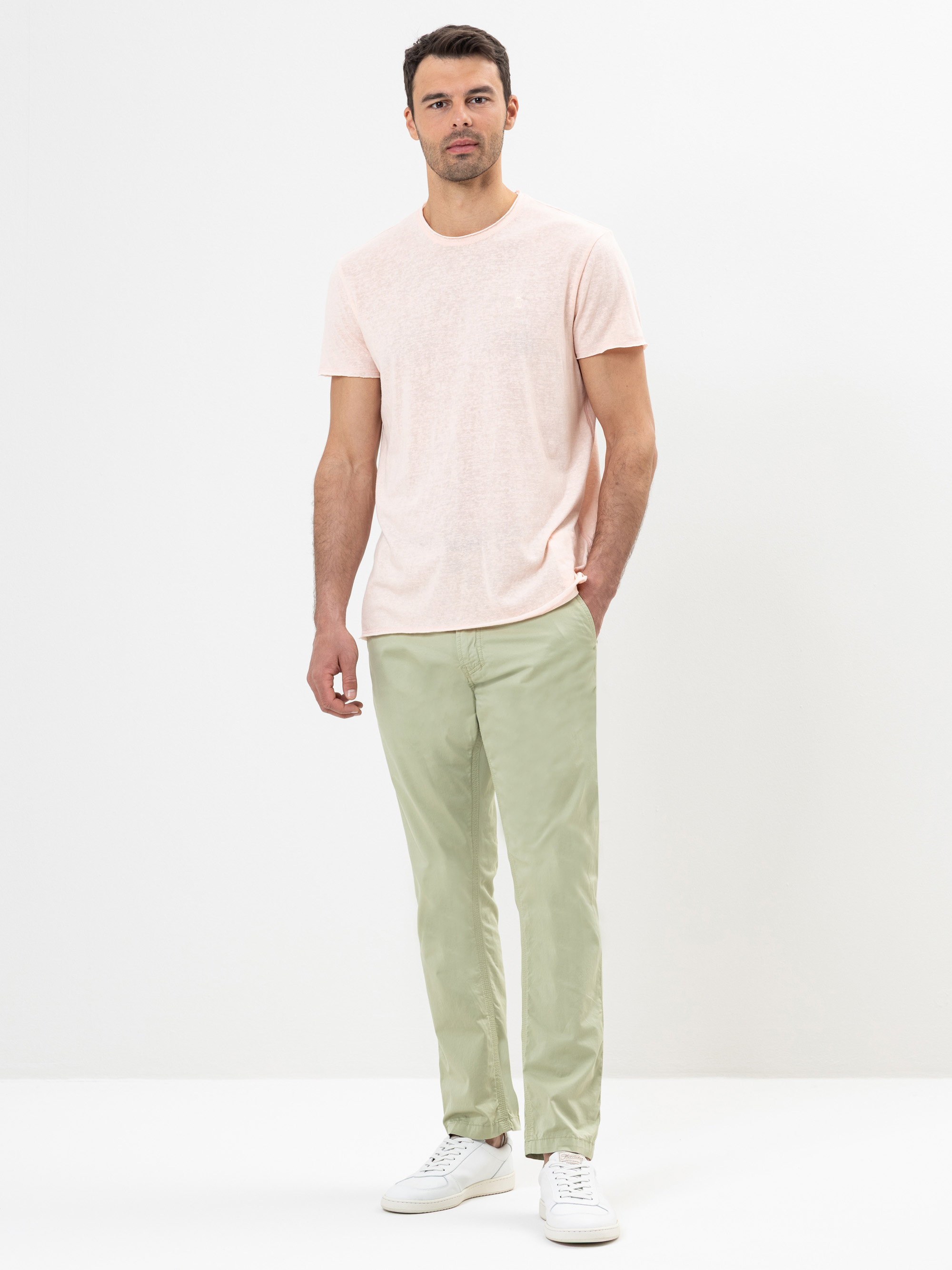 Ein Mann steht vor einem schlichten weißen Hintergrund und trägt ein hellrosa kurzärmeliges T-Shirt, eine hellgrüne Hose und weiße Turnschuhe. Seine linke Hand steckt in der Hosentasche und er blickt mit neutralem Blick geradeaus.