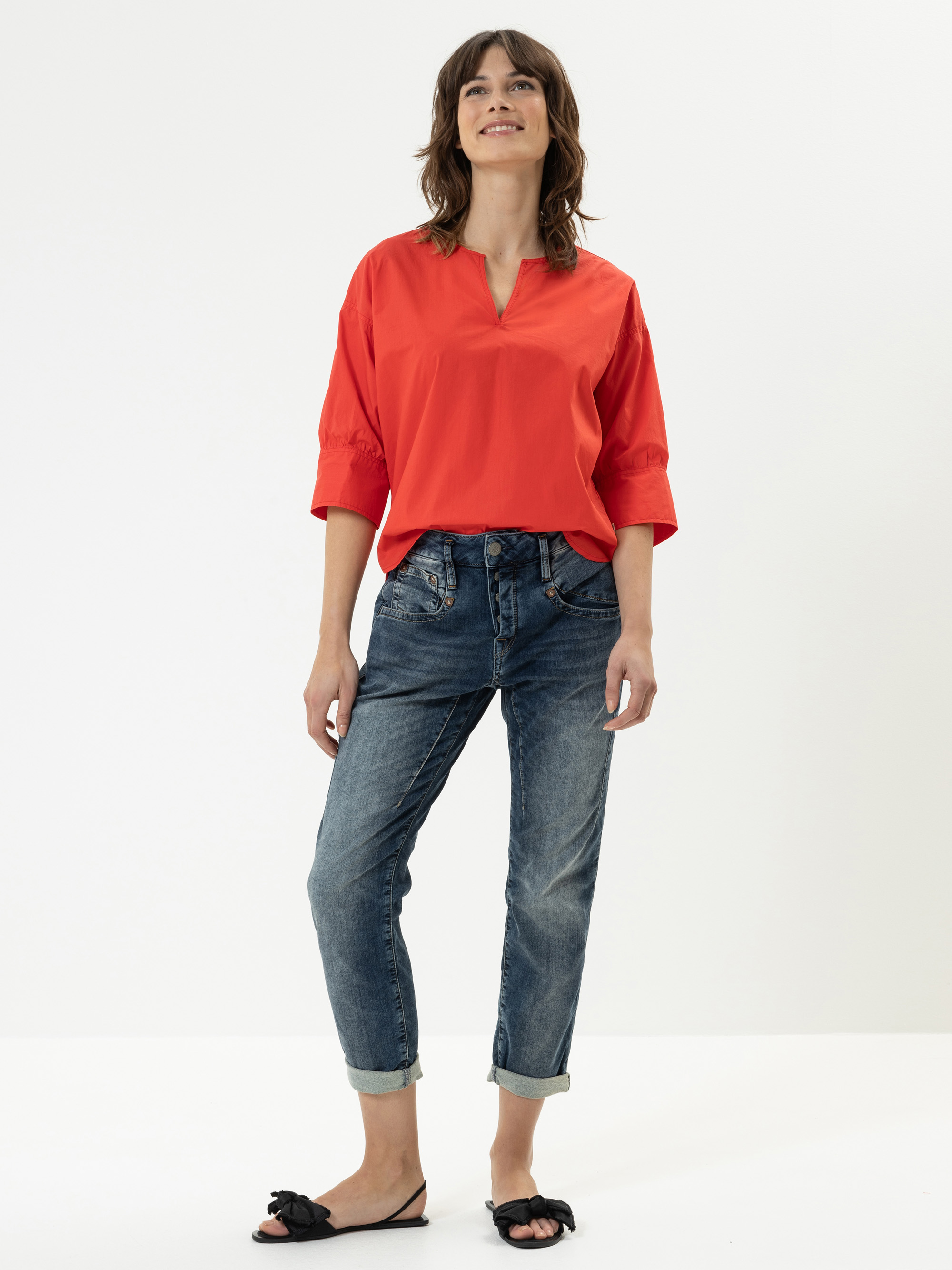 Eine Frau steht lächelnd vor einem schlichten weißen Hintergrund. Sie trägt eine leuchtend rote Bluse mit Dreiviertelärmeln, blaue, hochgekrempelte Jeans und schwarze Gleitsandalen mit einer Schleife.