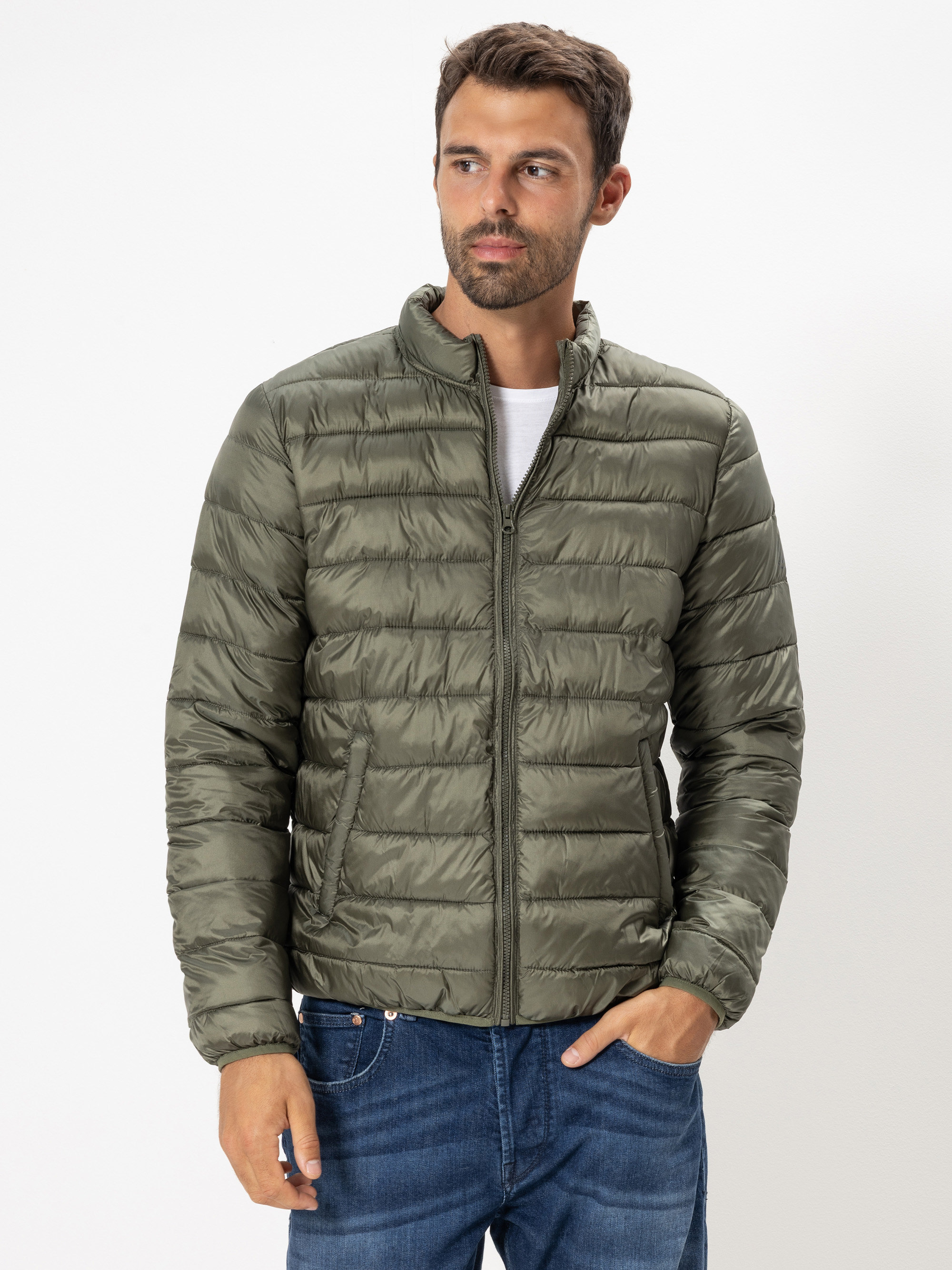 Ein Mann in einer hellen olivgrünen Steppjacke und blauen Jeans steht vor einem schlichten weißen Hintergrund, eine Hand in der Tasche, und schaut leicht zur Seite.