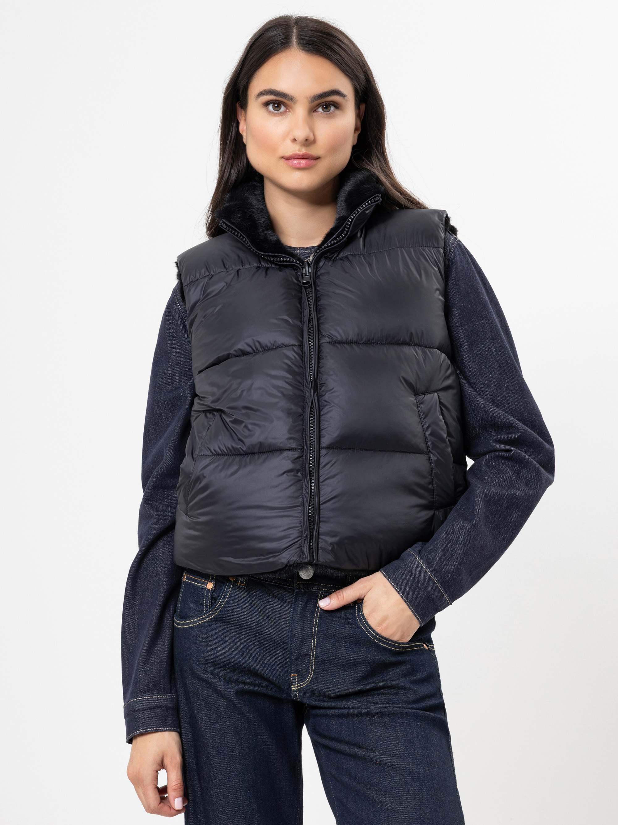 Eine Frau mit langen dunklen Haaren trägt eine schwarze Pufferweste über einer dunklen Jeansjacke und Jeans. Sie steht vor einem schlichten weißen Hintergrund und hat eine Hand in ihrer Tasche.