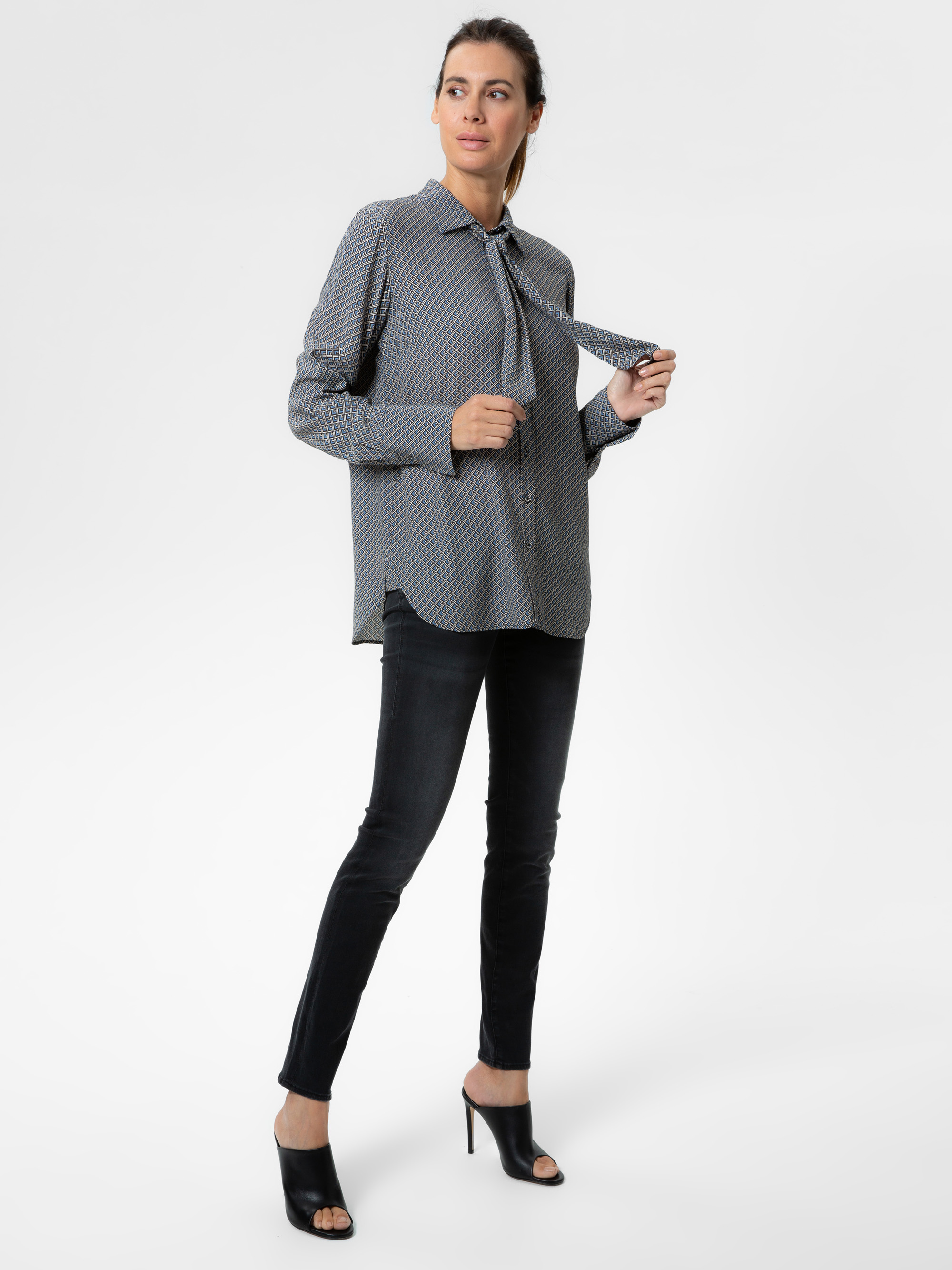 Eine Frau steht selbstbewusst in einer gemusterten langärmeligen Bluse mit Krawatte, schwarzen Röhrenjeans und schwarzen offenen Absätzen vor einem schlichten, hellen Hintergrund.