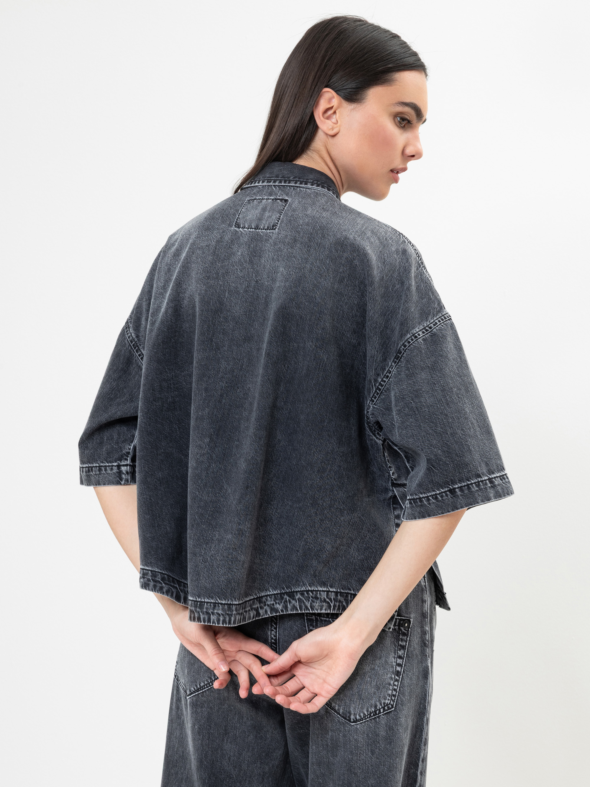 Eine Frau mit glattem dunklem Haar, die ein locker sitzendes, verblichenes schwarzes Jeanshemd und eine passende Hose trägt, steht mit dem Rücken zur Kamera, die Hände hinter sich verschränkt. Der Hintergrund ist einfarbig weiß.