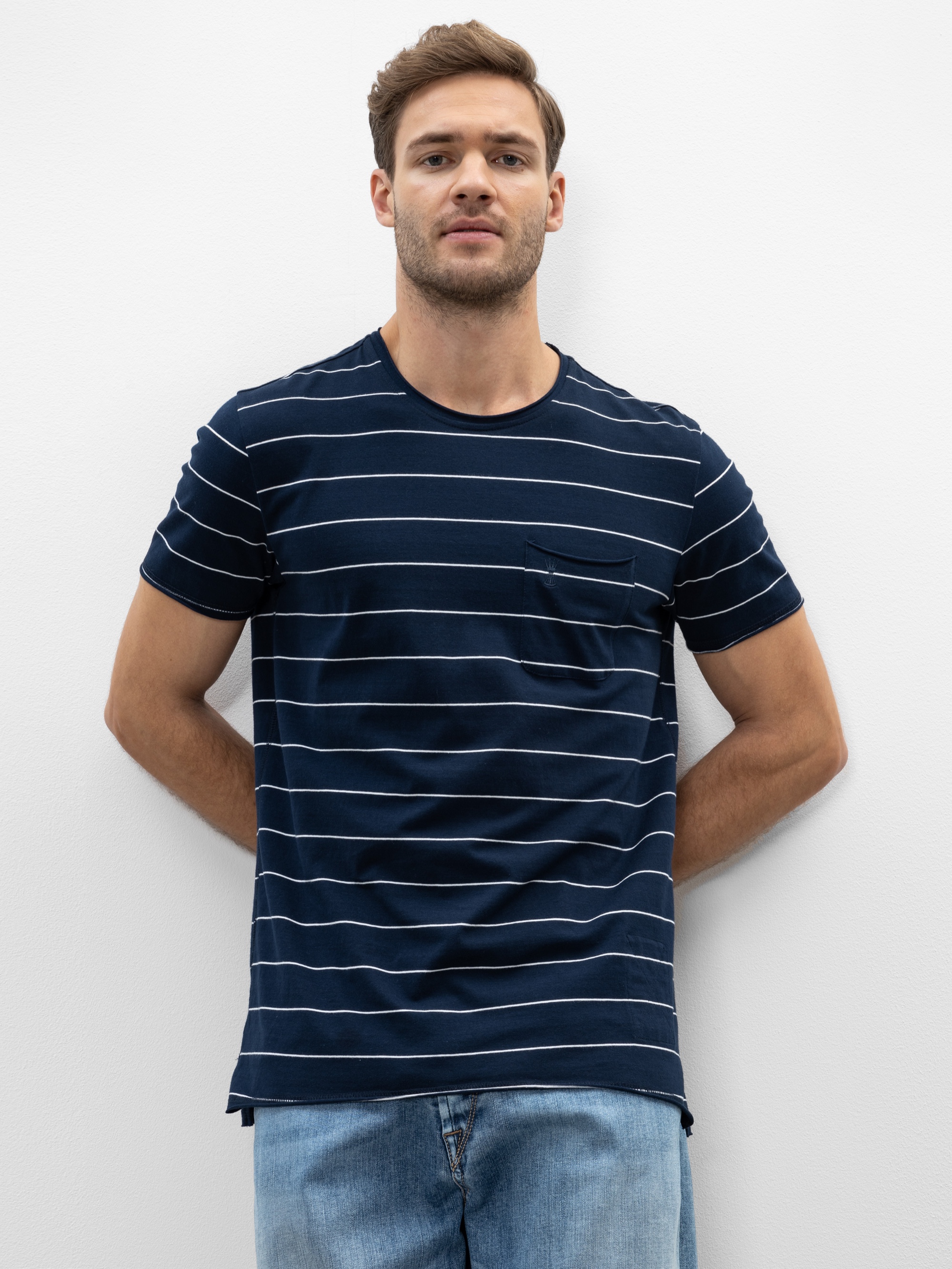 Ein Mann, der ein marineblaues T-Shirt mit weißen Querstreifen und hellblaue Jeans trägt, steht vor einer schlichten weißen Wand und schaut mit neutralem Blick in die Kamera.
