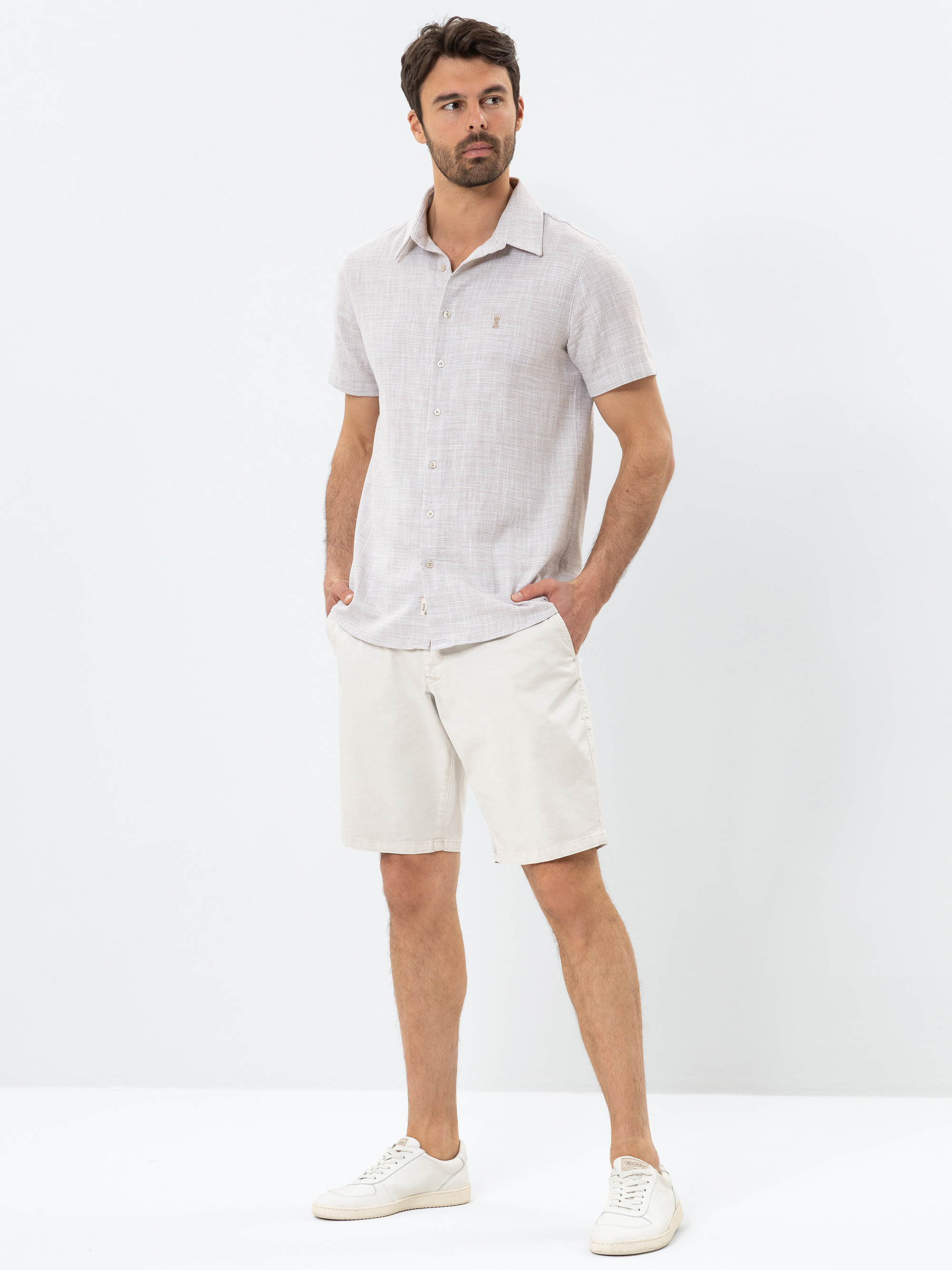 Ein Mann steht vor einem schlichten weißen Hintergrund, trägt ein hellgraues, kurzärmeliges Button-up-Hemd, hellbeige Shorts und weiße Turnschuhe, die Hände in den Taschen und einen neutralen Gesichtsausdruck.