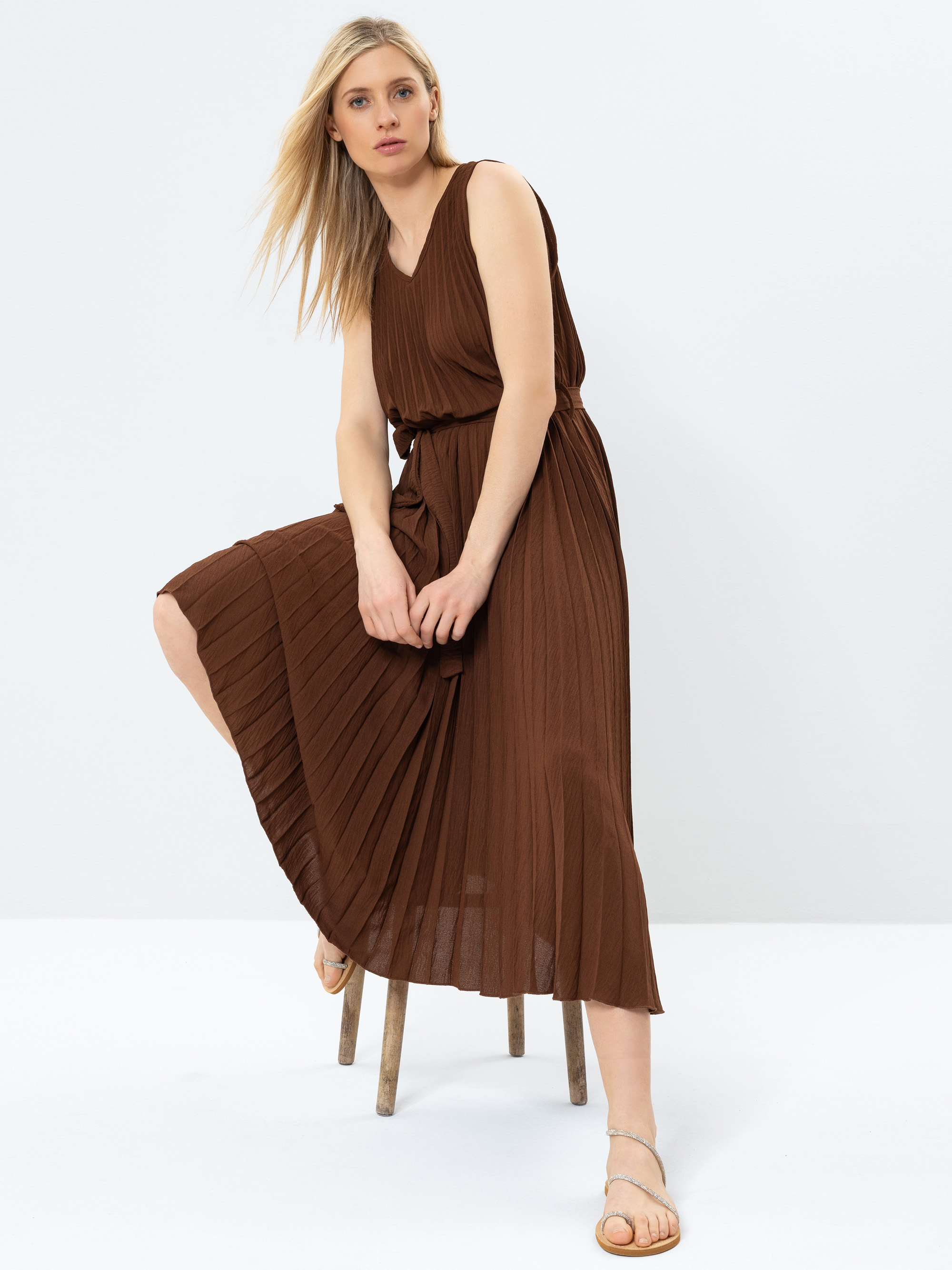Eine Frau mit langen blonden Haaren trägt ein ärmelloses, braunes, plissiertes Midikleid und beigefarbene Sandalen. Sie sitzt auf einem Holzschemel mit einem angehobenen Knie vor einem schlichten weißen Hintergrund.