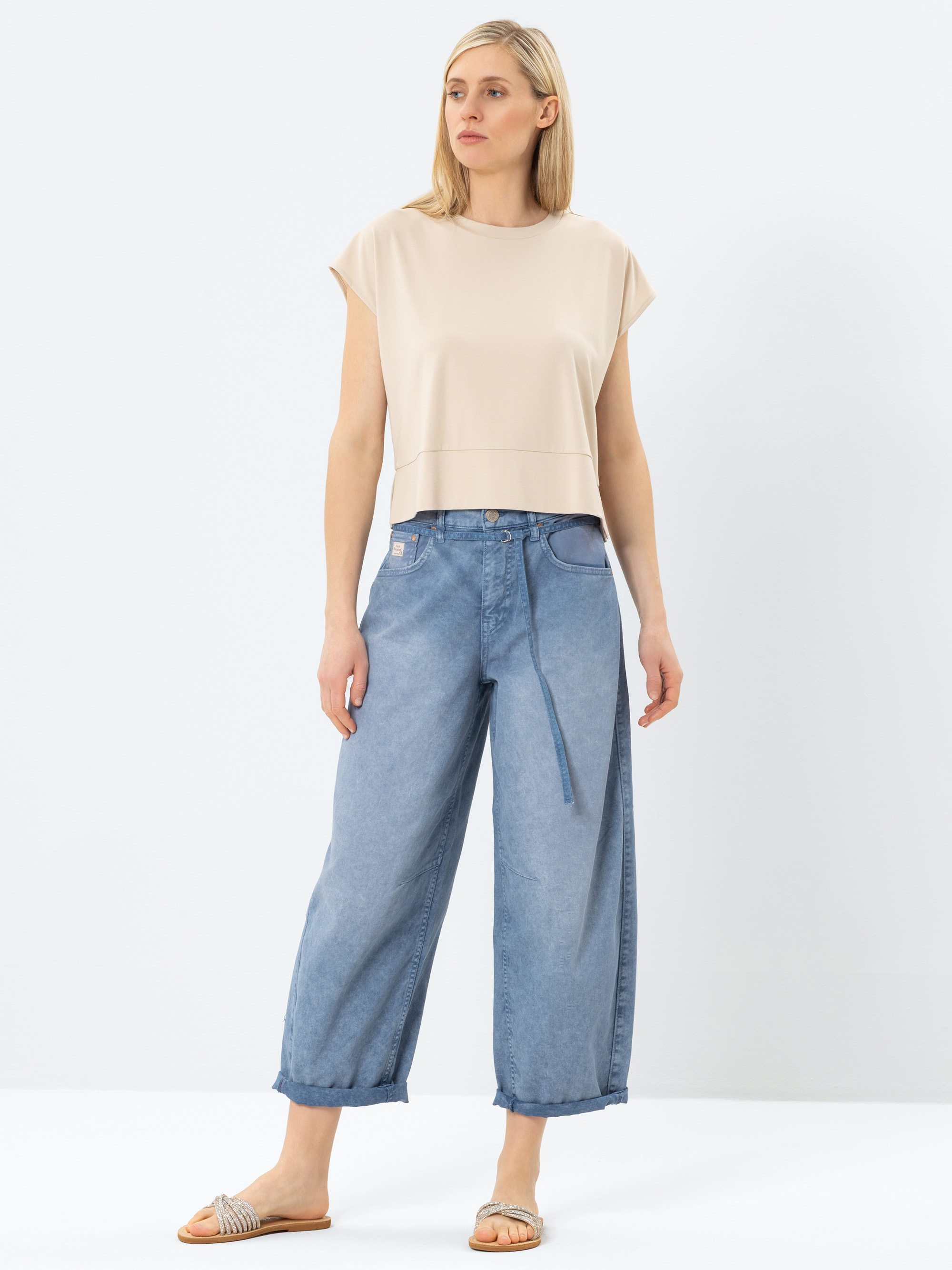 Eine Frau steht vor einem schlichten Hintergrund und trägt ein beigefarbenes kurzärmeliges Oberteil, locker sitzende hellblaue Jeans mit Gürtel und beigefarbene Sandalen. Sie hat glattes, hellblondes Haar und einen neutralen Gesichtsausdruck.