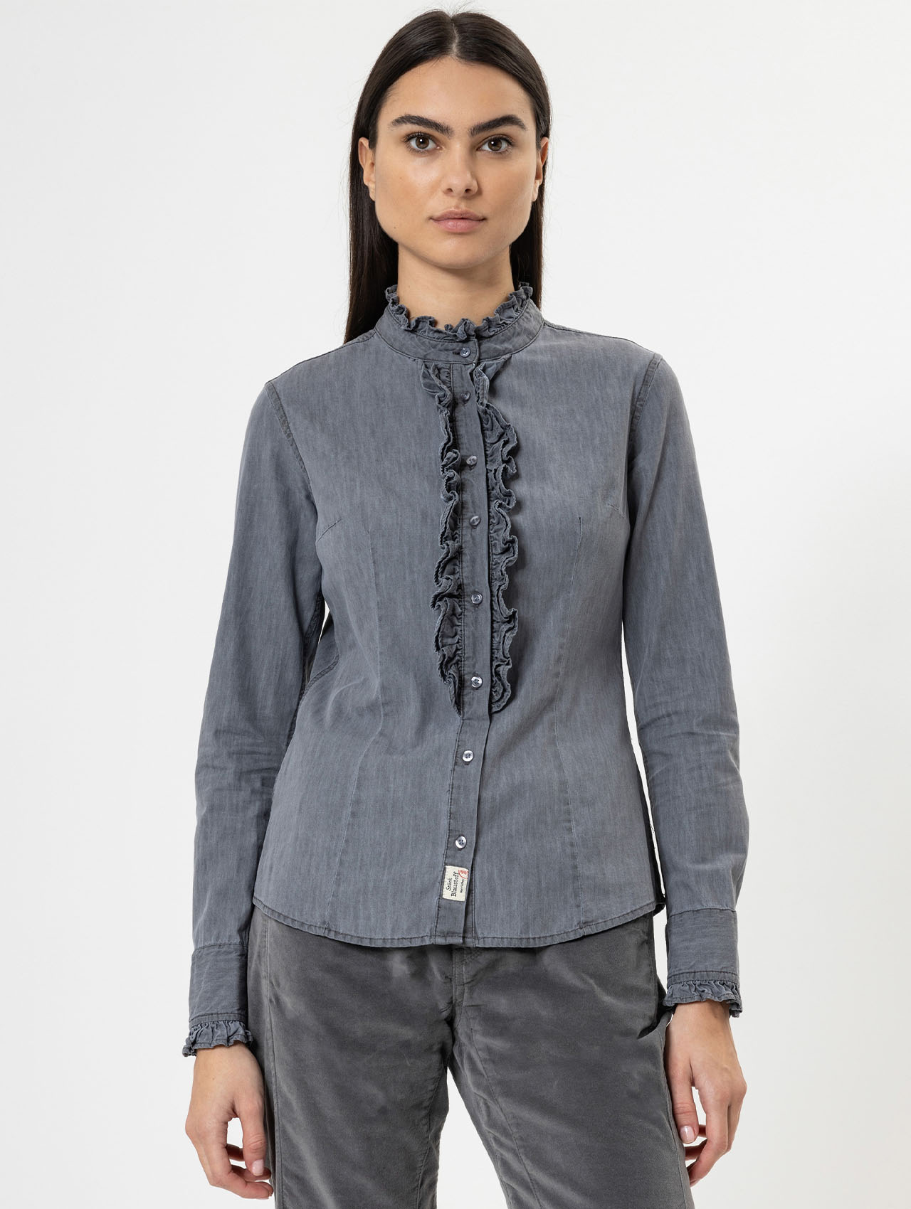 Eine Frau mit langen dunklen Haaren trägt eine graue, langärmelige Bluse mit Rüschen auf der Vorderseite und an den Manschetten. Sie steht vor einem schlichten weißen Hintergrund und blickt direkt in die Kamera.