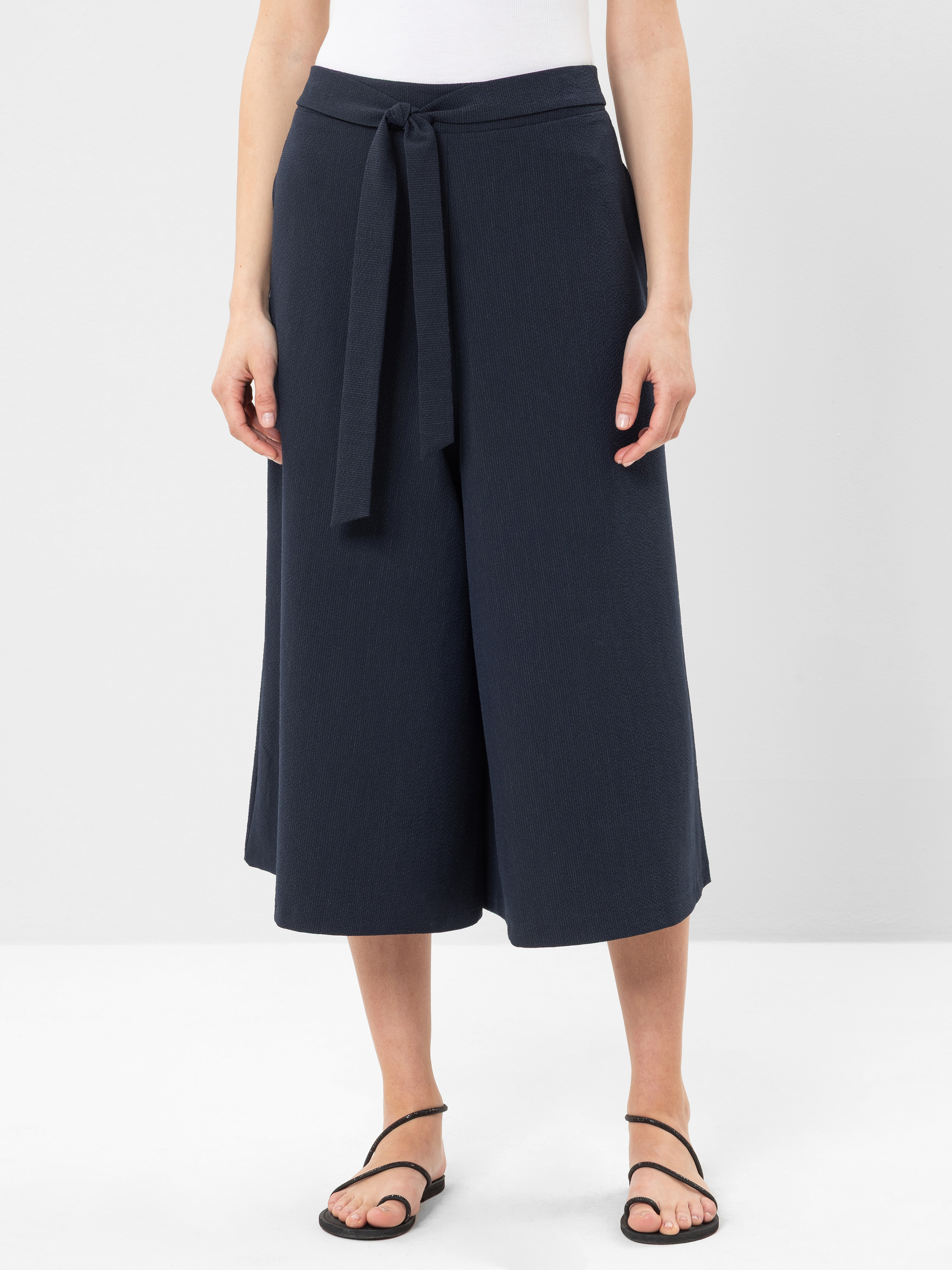 Eine Person, die marineblaue Culottes mit einem passenden Stoffgürtel, ein weißes ärmelloses Oberteil und schwarze Riemchensandalen trägt, steht vor einem schlichten weißen Hintergrund.