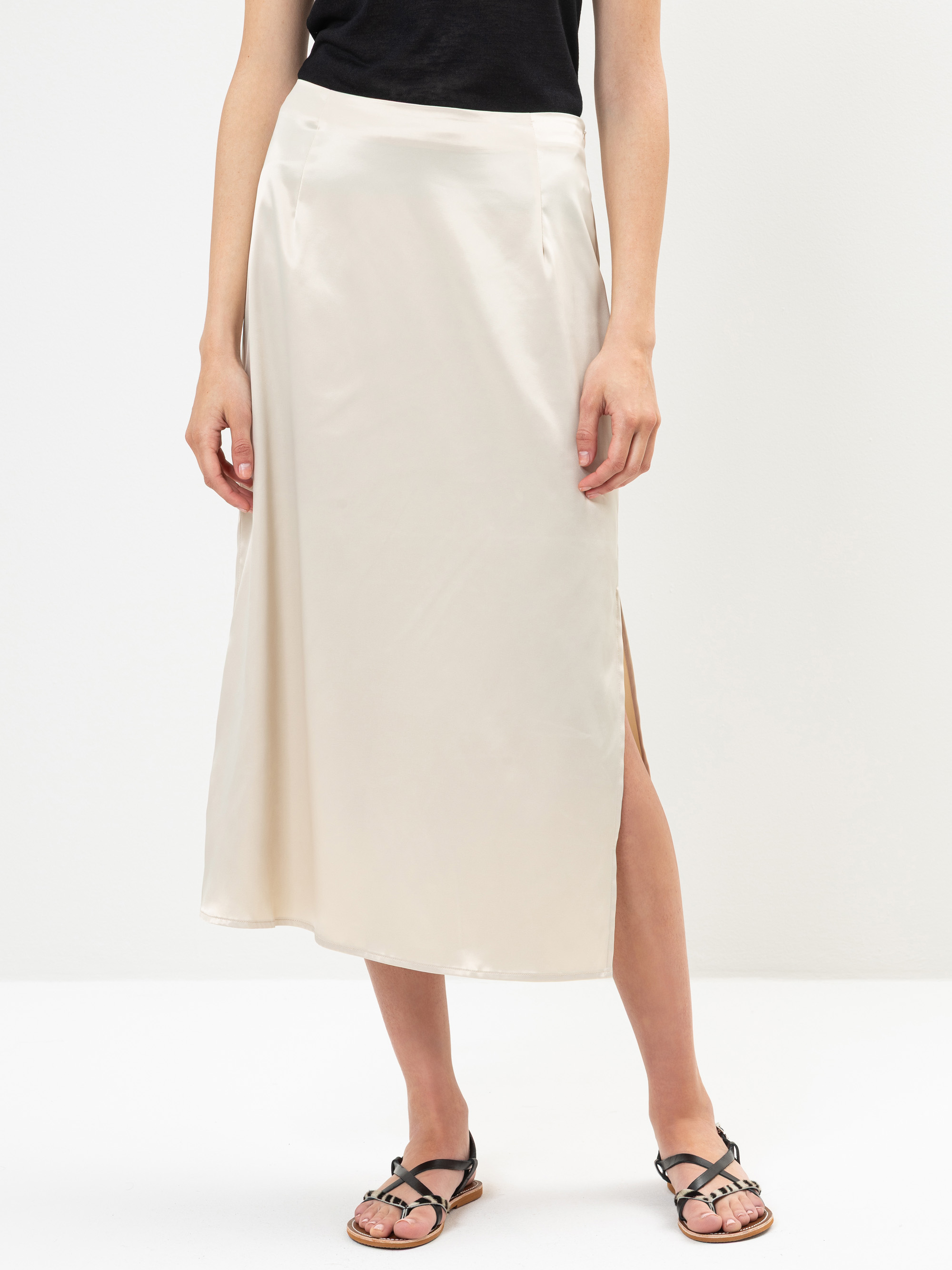Eine Person, die ein schwarzes Oberteil und einen cremefarbenen Midirock aus Satin mit Seitenschlitz trägt, gepaart mit schwarzen Riemchensandalen, steht vor einem schlichten weißen Hintergrund.