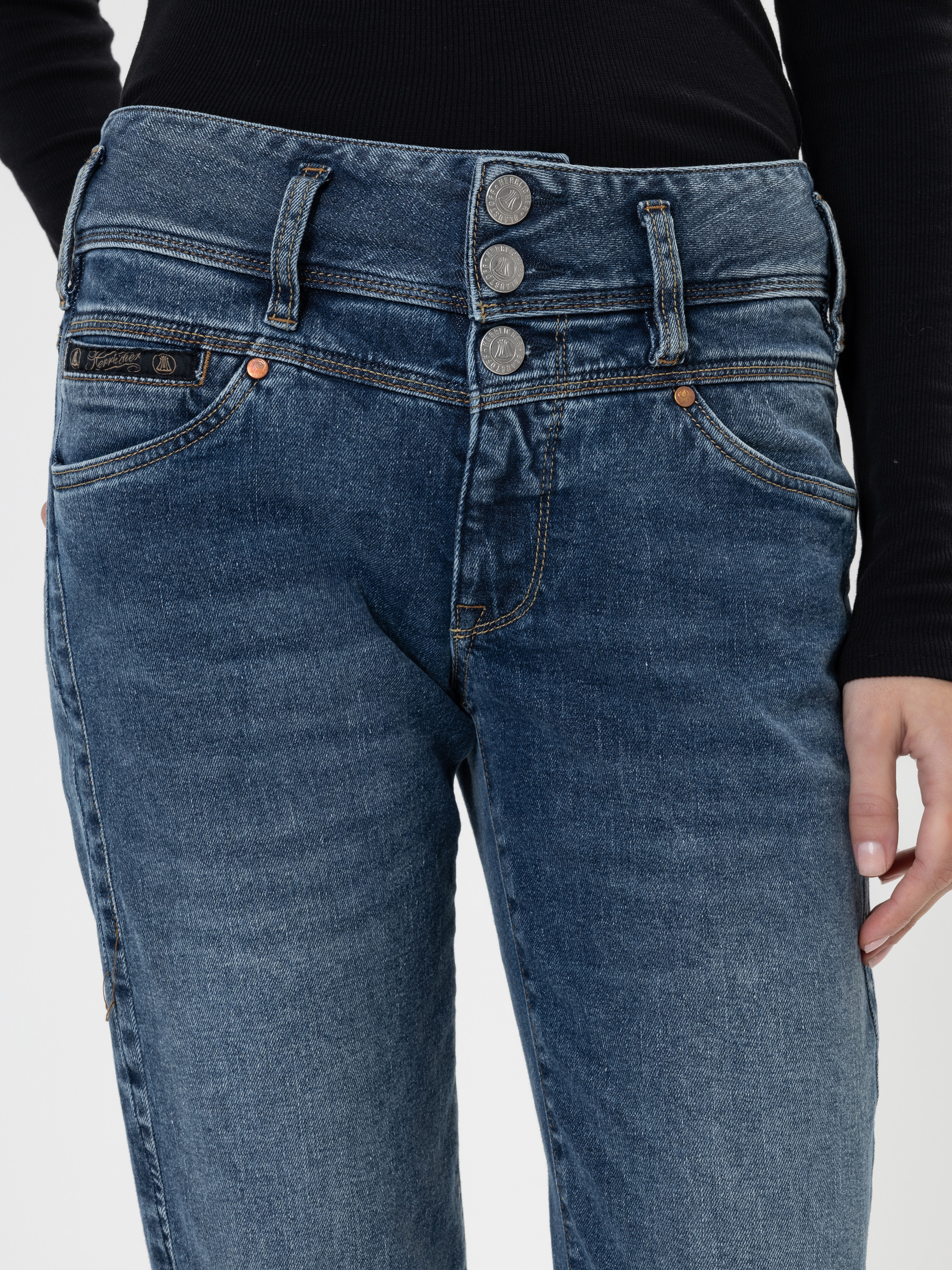 Eine Person trägt eine blaue Jeans mit hoher Taille und drei sichtbaren Knöpfen und ein schwarzes langärmeliges Oberteil, das von der Taille bis zur Mitte des Oberschenkels gezeigt wird.