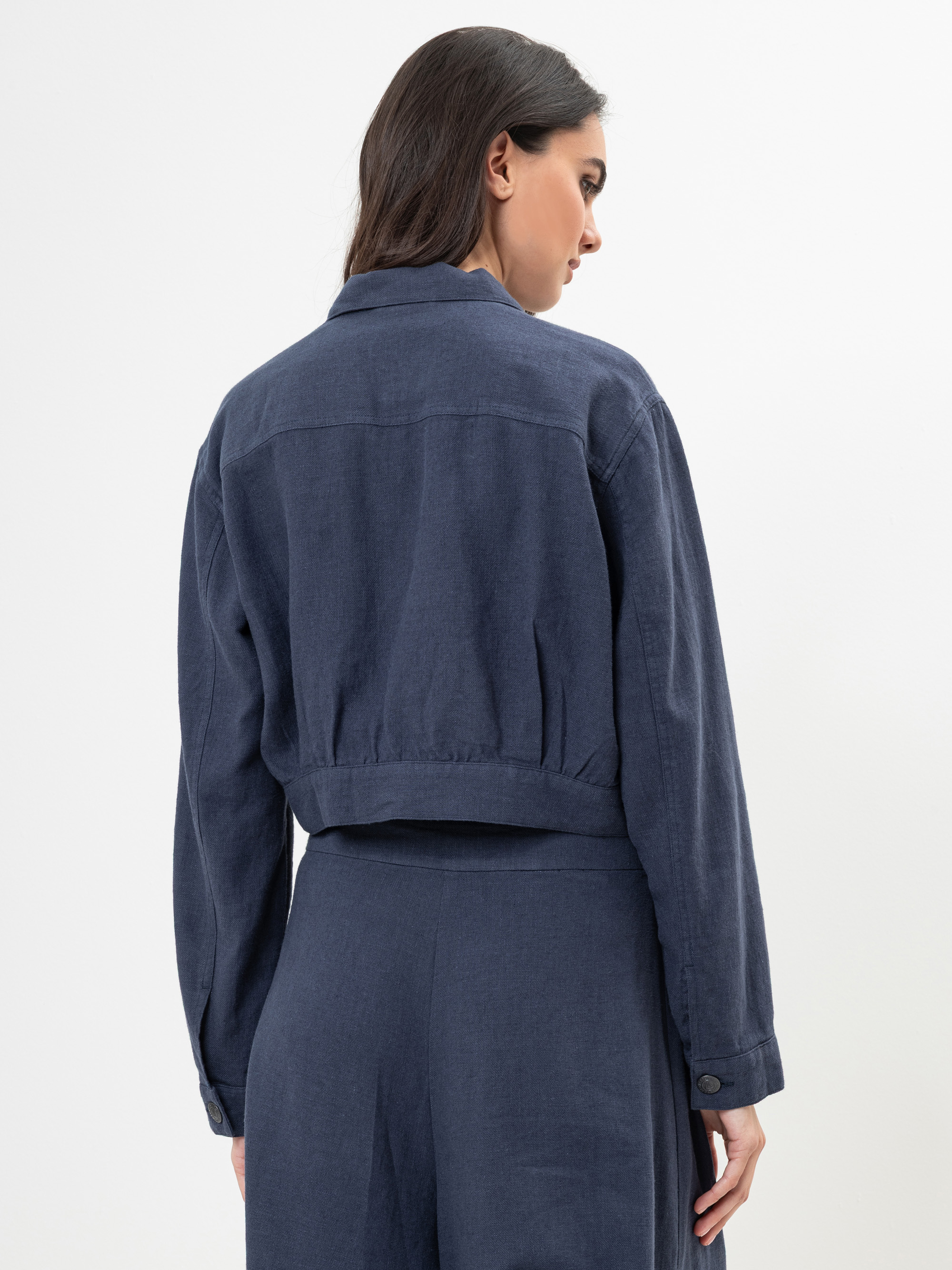 Eine Frau mit langen dunklen Haaren steht mit dem Gesicht zur Wand und trägt eine dunkelblaue Jacke mit Kragen und eine dazu passende Hose mit hoher Taille vor einem schlichten weißen Hintergrund.