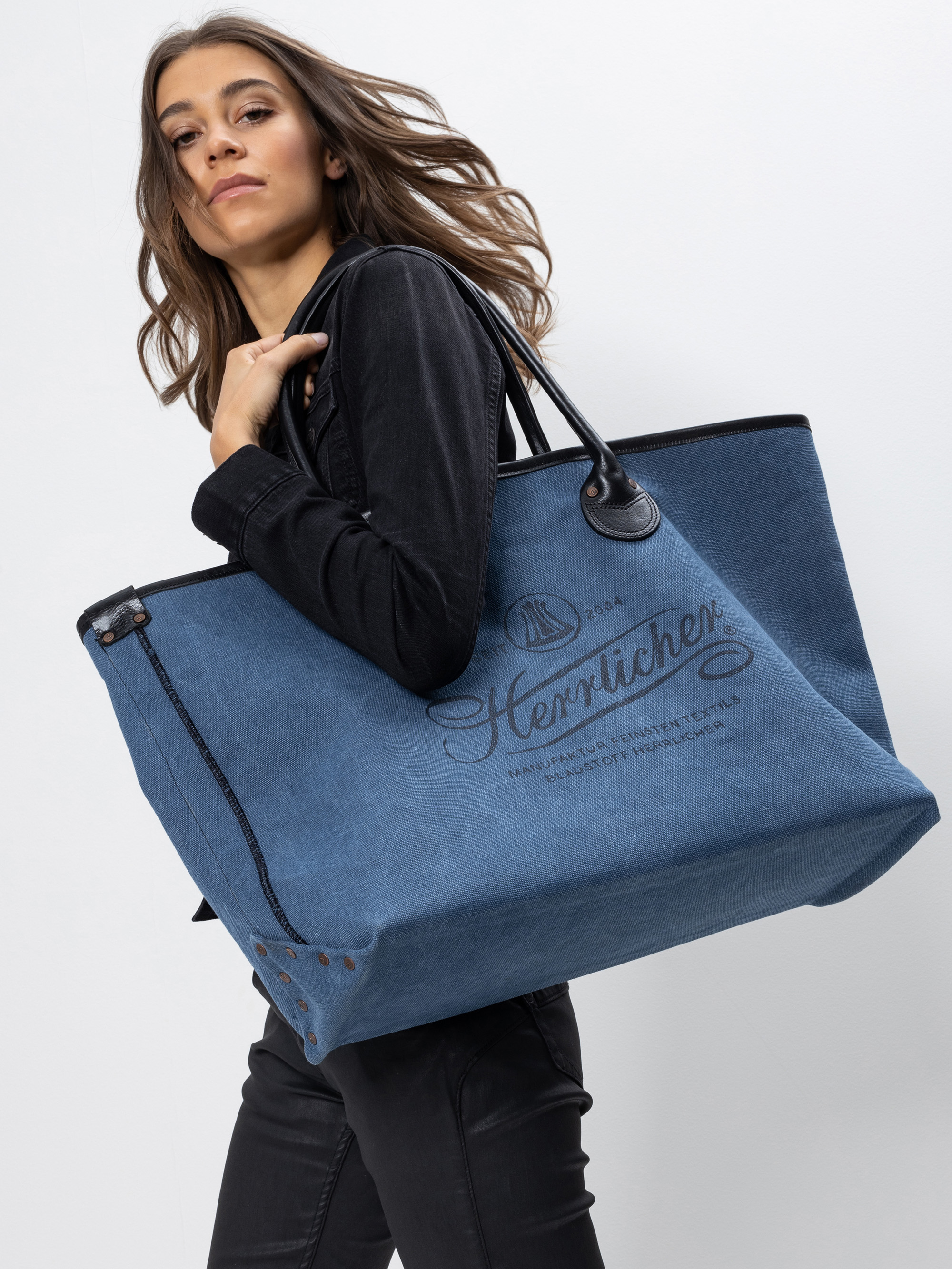 Eine Frau mit langen braunen Haaren und schwarzem Outfit trägt eine große blaue Tragetasche aus Jeansstoff mit schwarzen Henkeln und dekorativem Text auf der Vorderseite, die vor einem schlichten weißen Hintergrund steht.