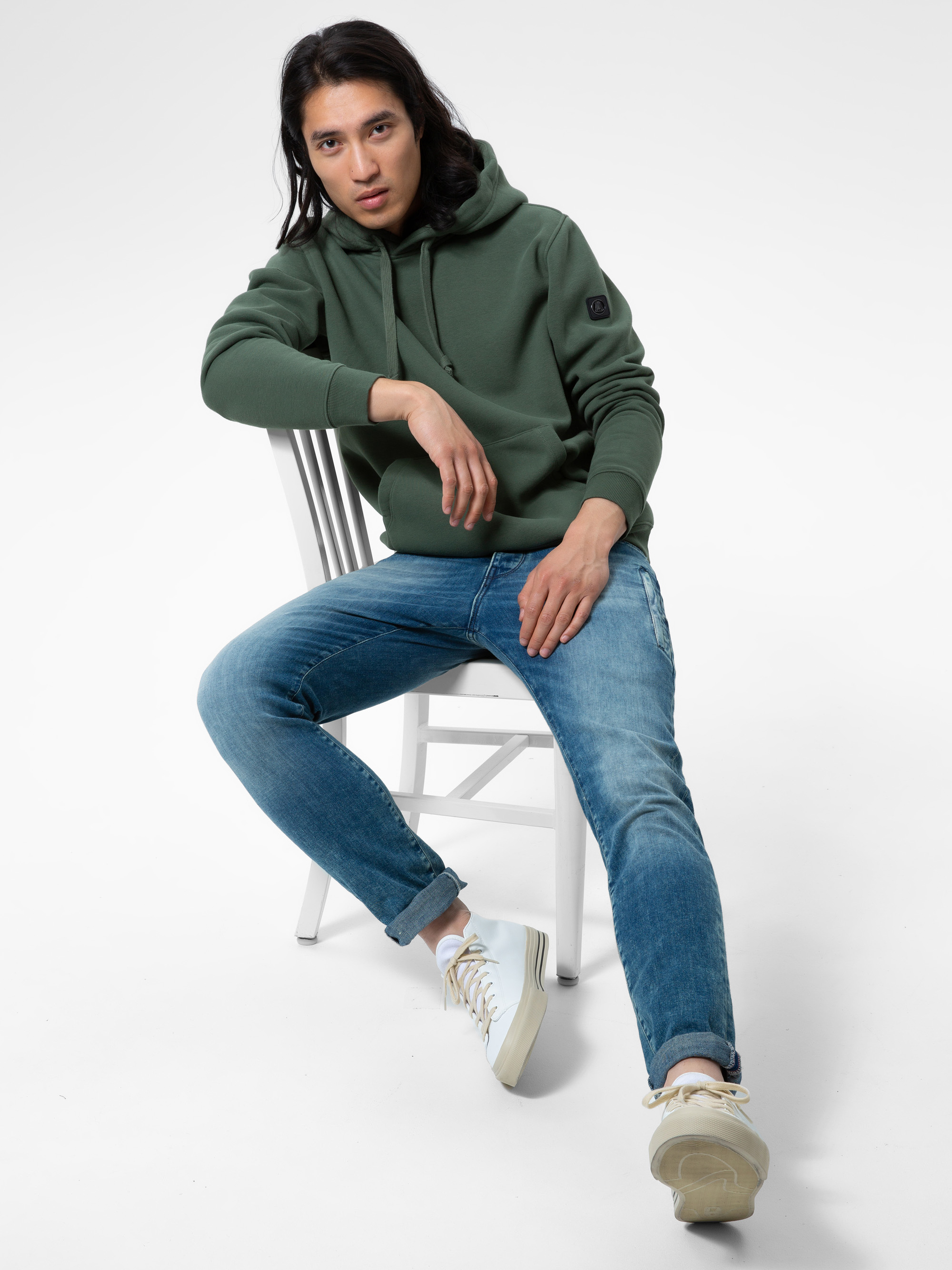 Eine Person mit langen dunklen Haaren sitzt seitlich auf einem weißen Stuhl, trägt einen grünen Kapuzenpullover, blaue Jeans und weiße Turnschuhe und blickt vor einem schlichten weißen Hintergrund direkt in die Kamera.