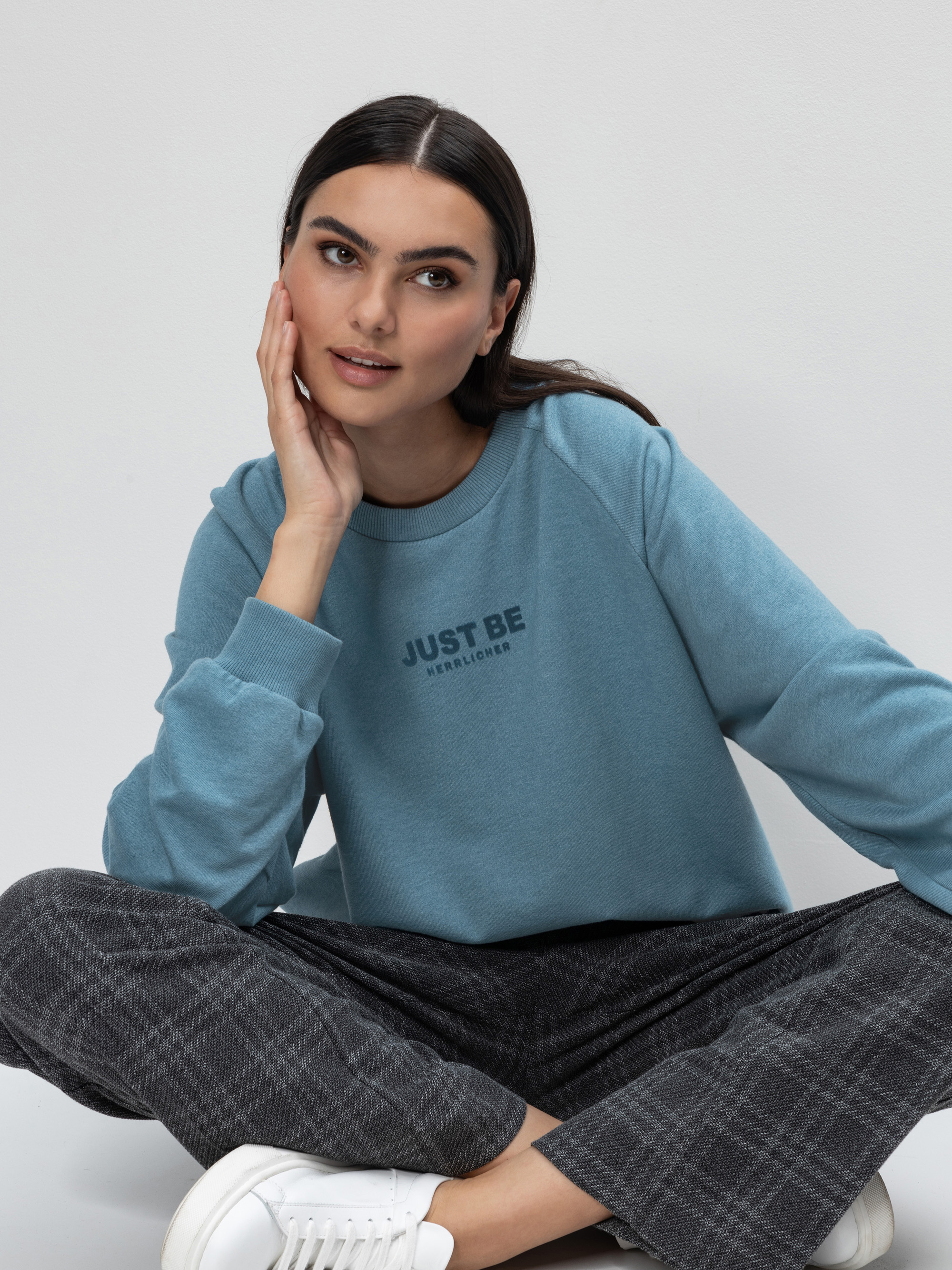 Eine Frau mit langen dunklen Haaren sitzt auf dem Boden, trägt ein hellblaues Sweatshirt mit der Aufschrift JUST BE, eine grau karierte Hose und weiße Turnschuhe, stützt ihr Kinn auf die Hand und schaut zur Seite.