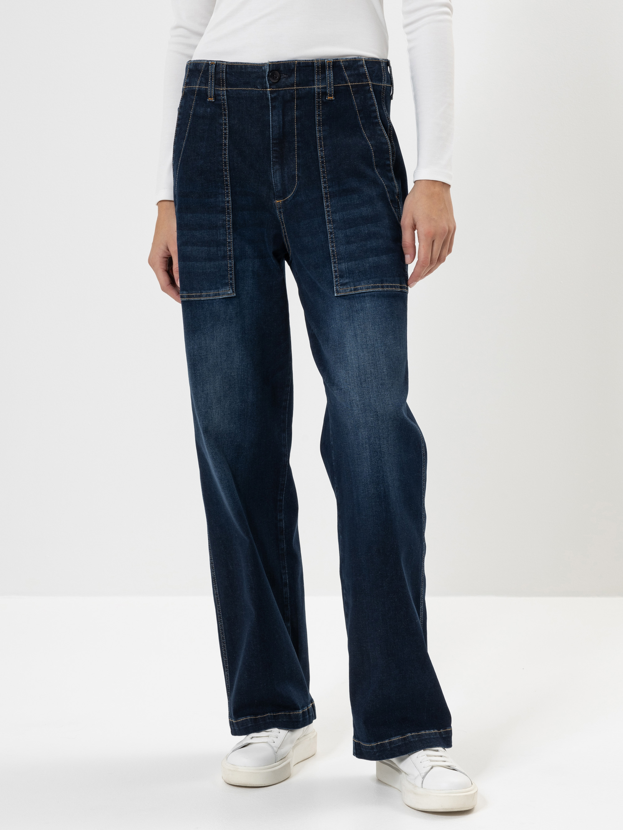 Person, die eine dunkle Jeans mit weitem Bein und großen Taschen auf der Vorderseite, ein weißes langärmeliges Oberteil und weiße Turnschuhe trägt und vor einem schlichten weißen Hintergrund steht. Der Kopf ist auf dem Bild nicht zu sehen.