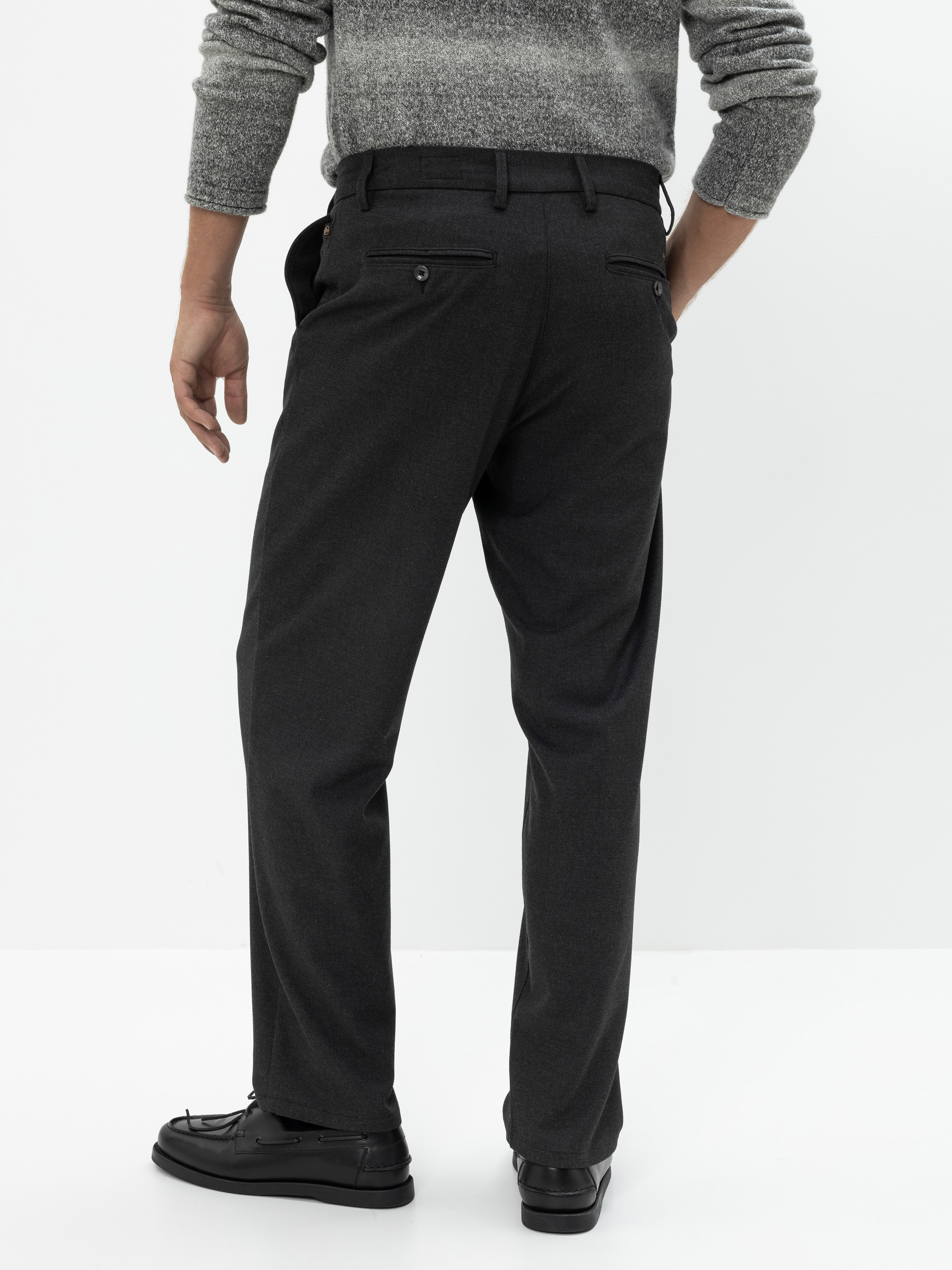 Ein Mann steht mit dem Gesicht nach hinten, trägt eine dunkelgraue Hose, einen hellgrauen Pullover und schwarze Schuhe vor einem schlichten weißen Hintergrund.