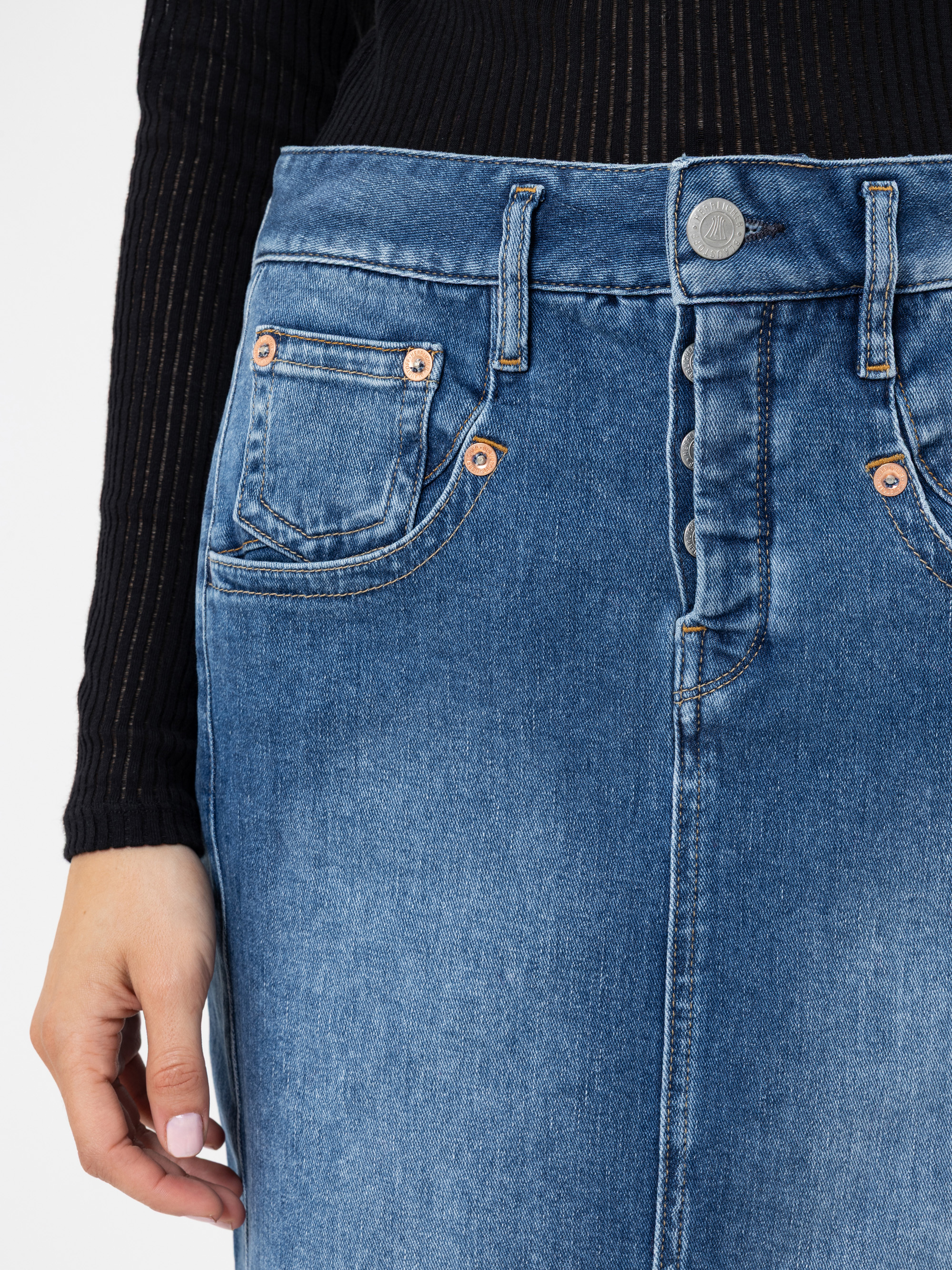 Eine Person trägt ein schwarzes, langärmeliges, geripptes Oberteil und einen blauen Jeansrock mit sichtbaren Knöpfen und orangefarbenen Nieten, wobei die Vordertasche und die Gürtelschlaufen zu sehen sind. Nur der untere Teil des Oberkörpers und die