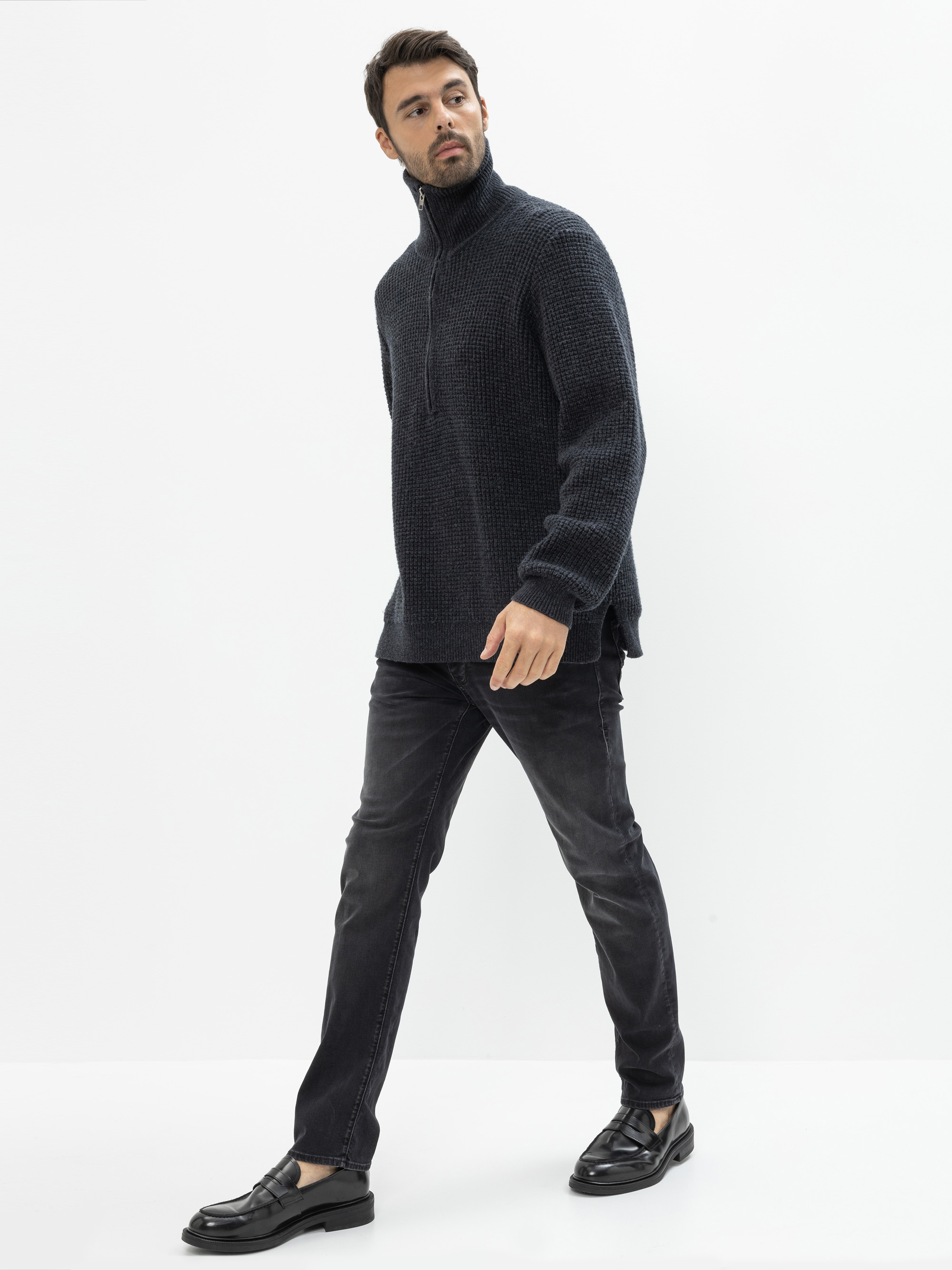 Ein Mann mit kurzen dunklen Haaren und einem Bart trägt einen strukturierten dunklen Pullover mit Reißverschluss, schwarze Jeans und schwarze Halbschuhe und geht selbstbewusst vor einem schlichten weißen Hintergrund.