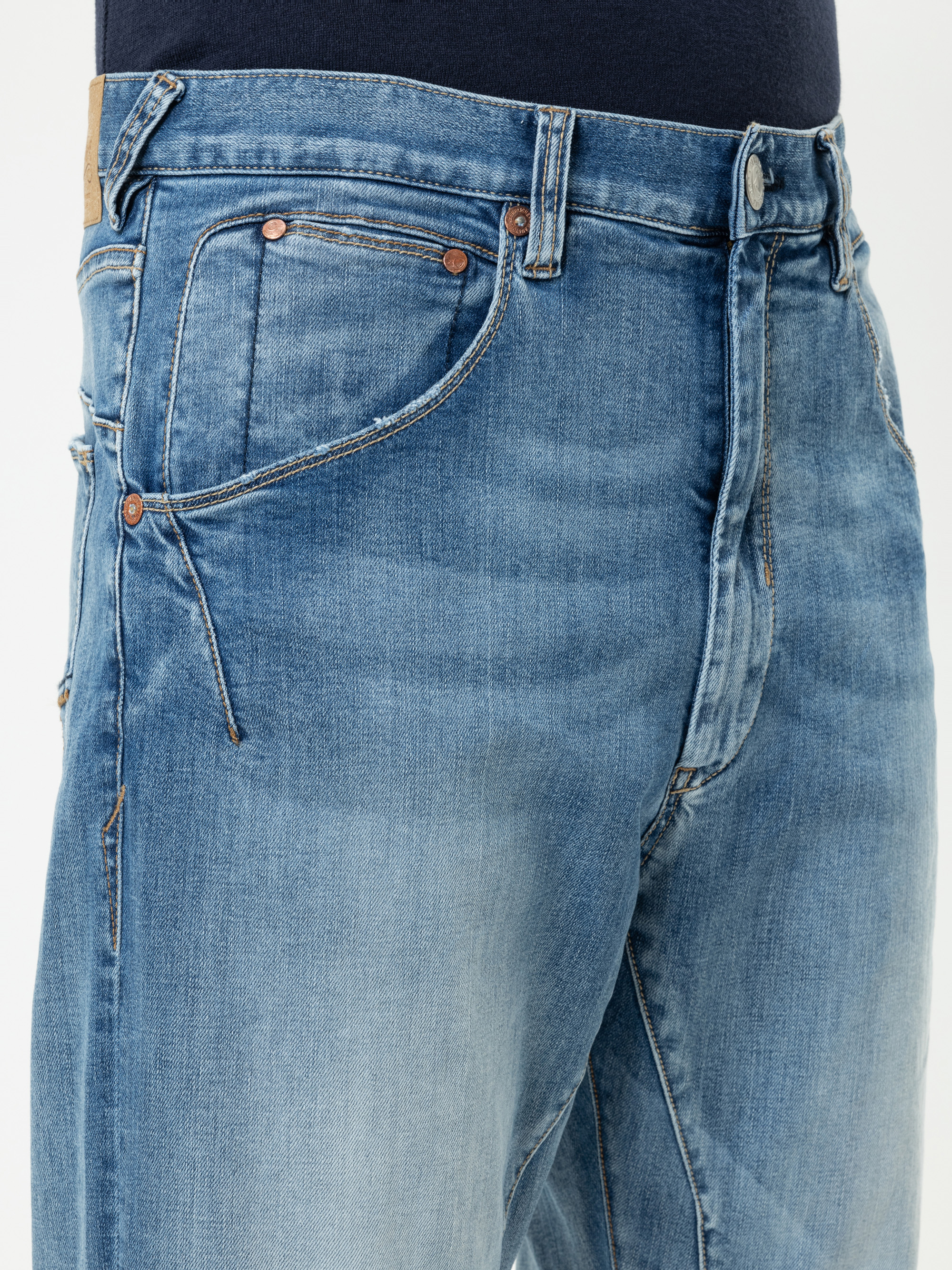 Nahaufnahme einer Person, die eine blaue Jeans mit verblasster Waschung, Details an der Vordertasche sowie einem sichtbaren Knopf und Bund vor einem einfarbigen Hintergrund trägt.