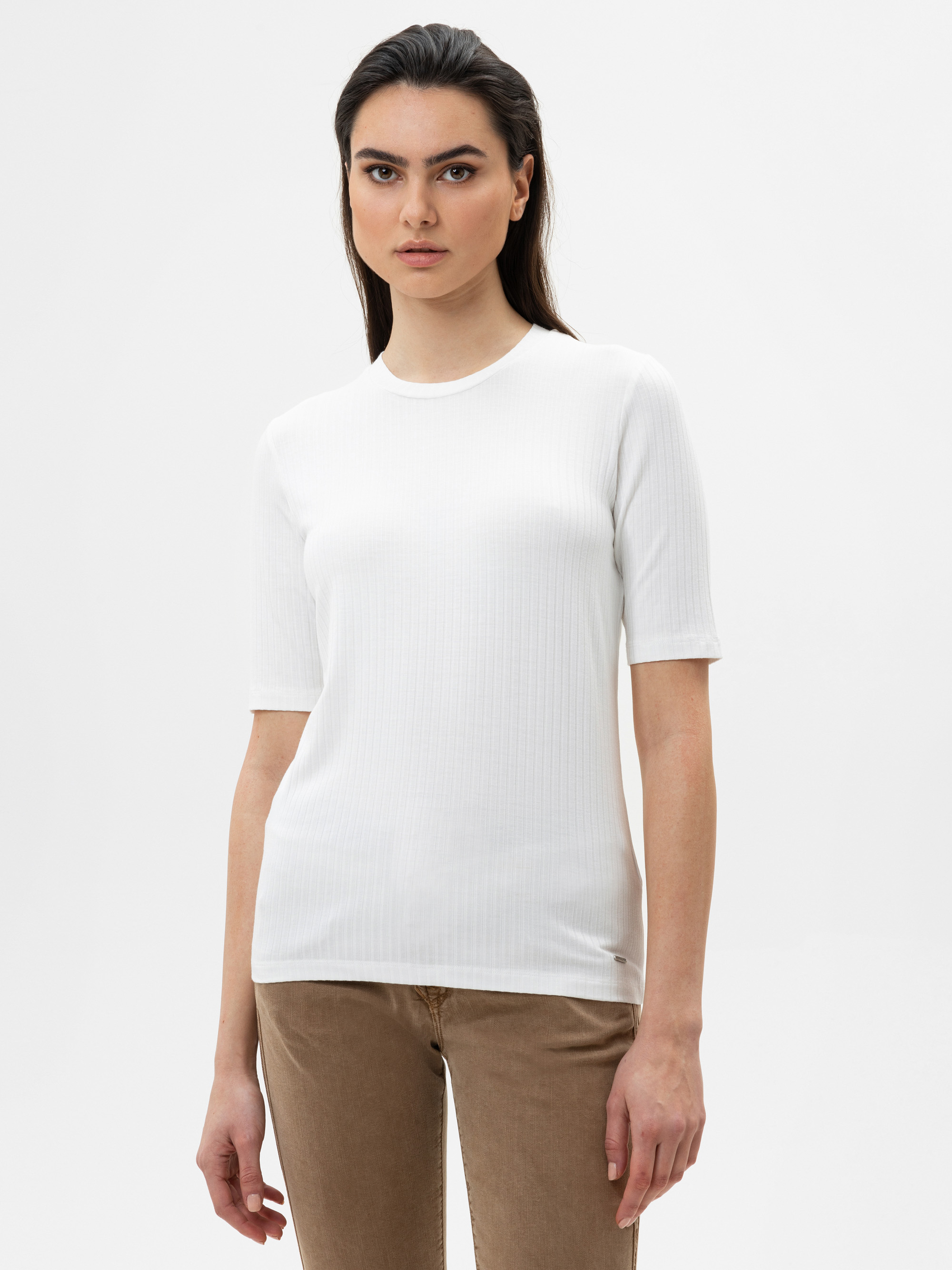 Eine Frau mit langen dunklen Haaren, die ein weißes, kurzärmeliges, geripptes Hemd und eine hellbraune Hose trägt, steht vor einem schlichten weißen Hintergrund und schaut mit neutralem Blick direkt in die Kamera.