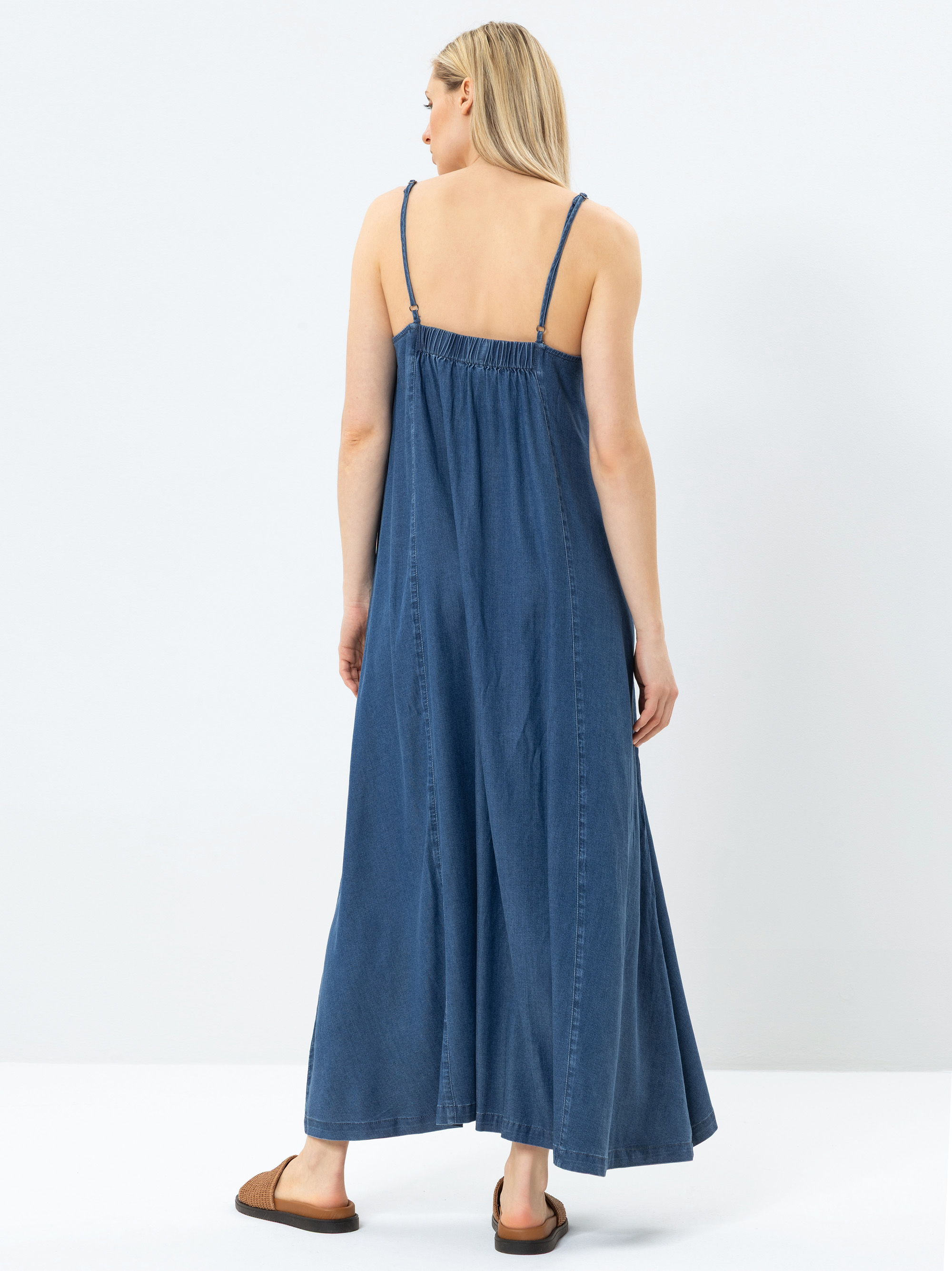 Eine Frau mit langen blonden Haaren trägt ein ärmelloses, blaues Jeans-Maxikleid mit dünnen Trägern und braune Sandalen. Sie steht vor einem schlichten weißen Hintergrund und blickt von der Kamera weg.