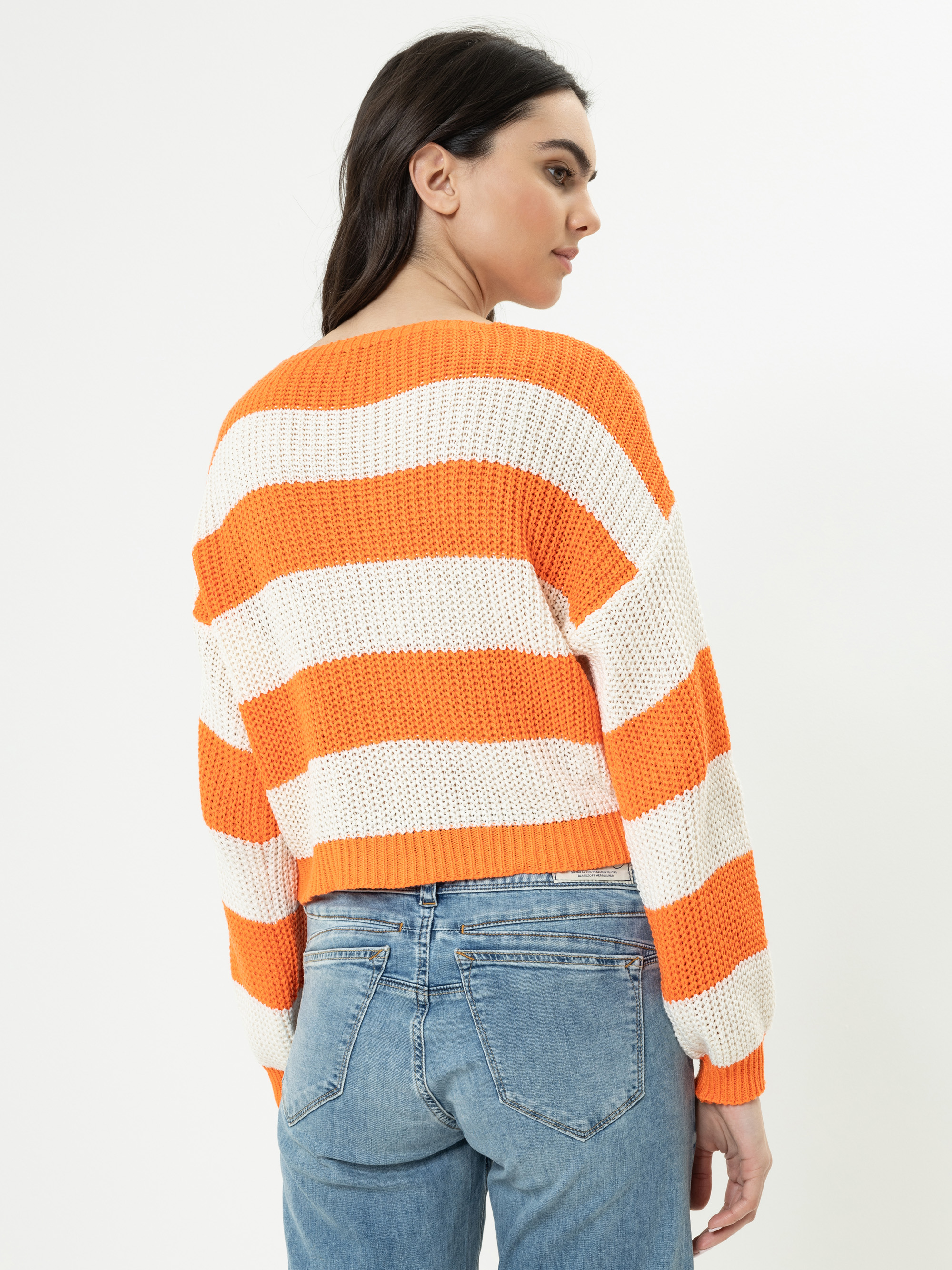 Eine Frau mit langen dunklen Haaren, die einen dicken Strickpulli mit breiten orangefarbenen und weißen Querstreifen und eine blaue Jeans trägt, steht mit dem Gesicht zur Wand und blickt über ihre Schulter auf einen unifarbenen Hintergrund.