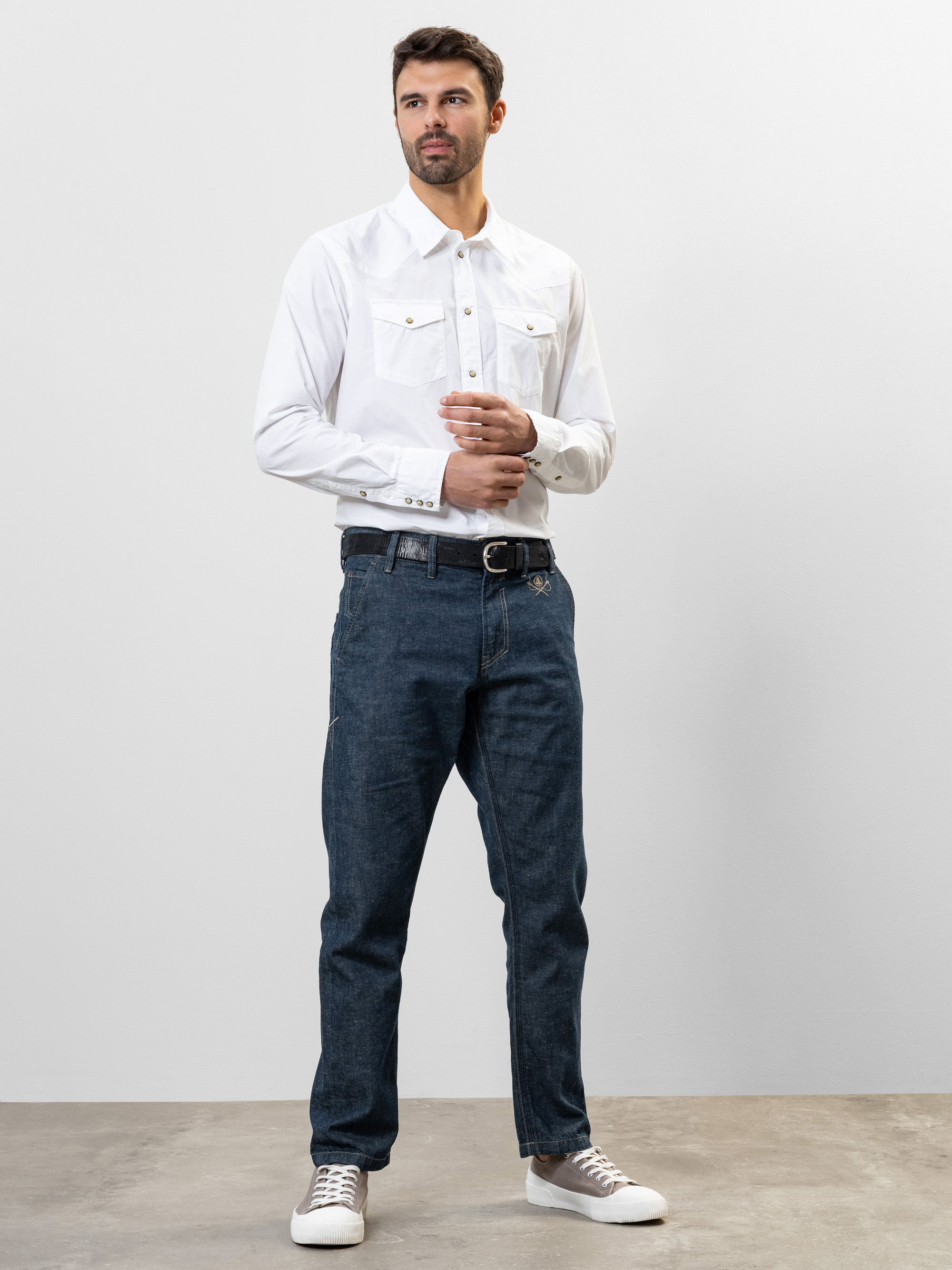 Ein Mann steht vor einem schlichten, hellen Hintergrund und trägt ein weißes Button-up-Hemd, blaue Jeans, einen schwarzen Gürtel und weiße Turnschuhe. Er hat kurzes dunkles Haar, einen Bart und die Hände locker vor sich verschränkt.