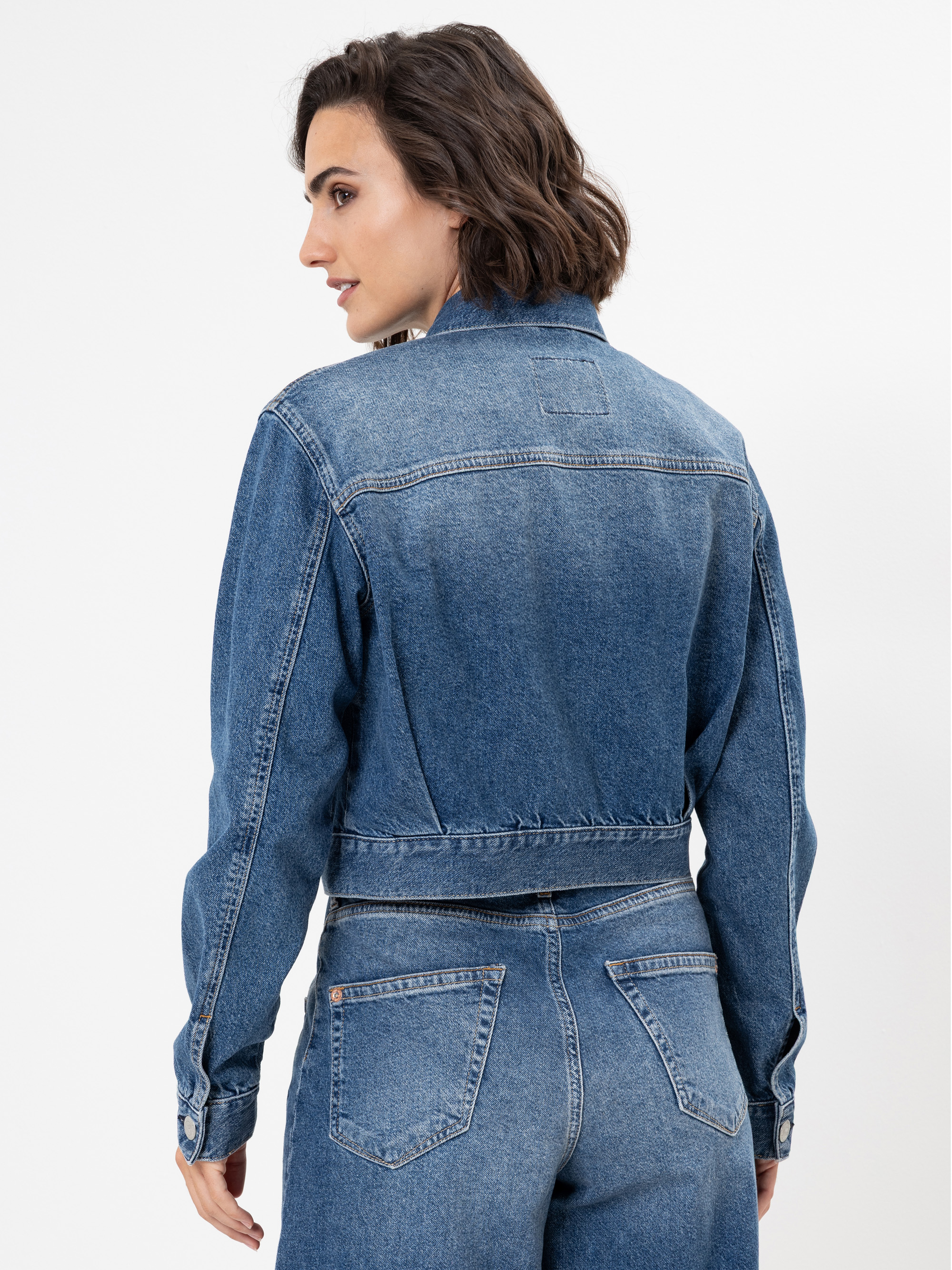 Eine Frau mit kurzen braunen Haaren ist von hinten zu sehen. Sie trägt eine blaue Jeansjacke und dazu passende Jeans mit hoher Taille. Sie steht vor einem schlichten weißen Hintergrund.