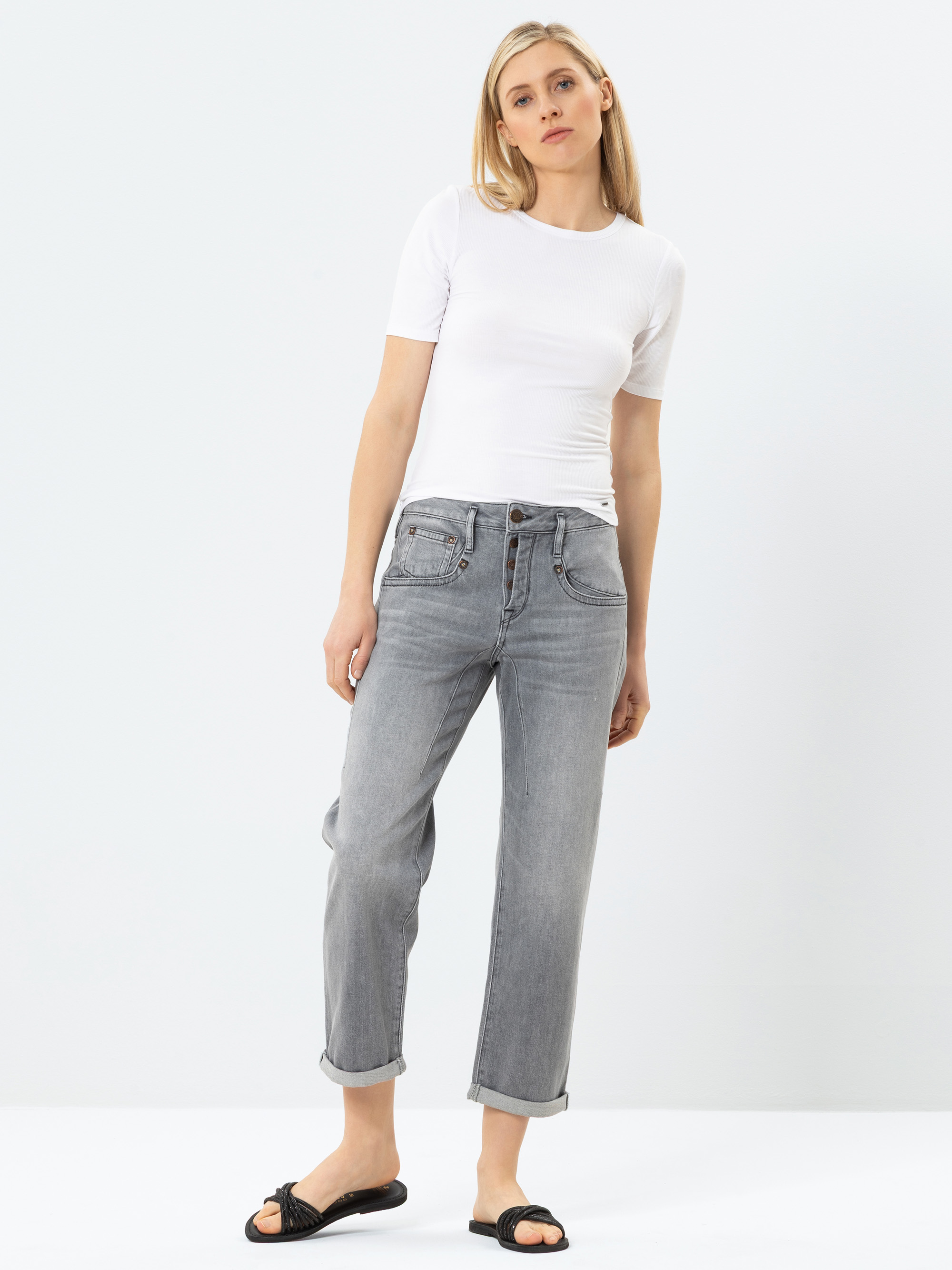 Eine Frau steht vor einem schlichten weißen Hintergrund und trägt ein kurzärmeliges weißes T-Shirt, hellgraue Jeans mit Bündchen und schwarze Slide-Sandalen. Sie hat blondes Haar und eine entspannte, lässige Pose.