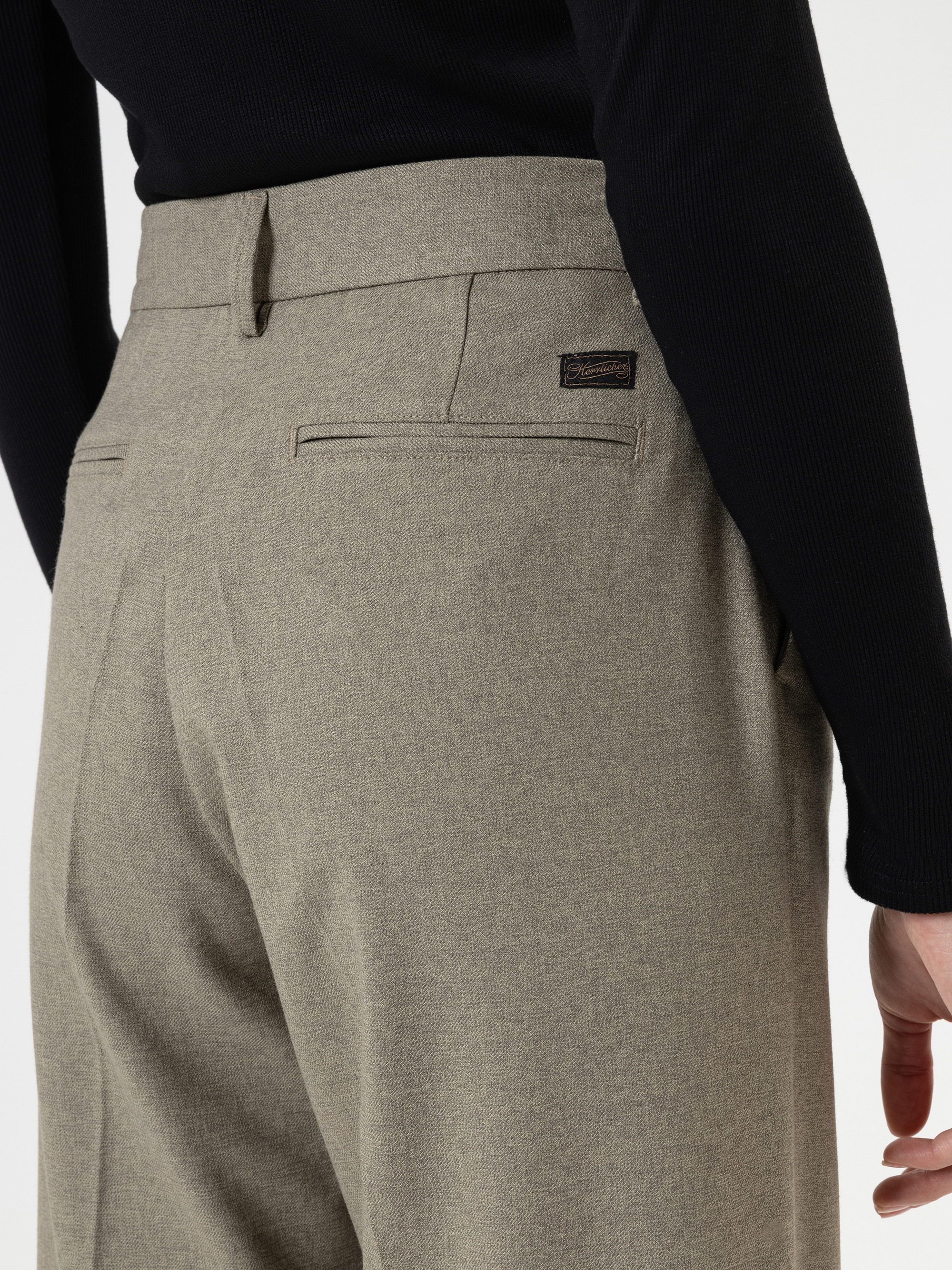 Eine Person, die eine hellbraune, maßgeschneiderte Hose und ein schwarzes, langärmeliges Oberteil trägt, wird von der Taille abwärts gezeigt; eine Hand liegt entspannt an ihrer Seite.