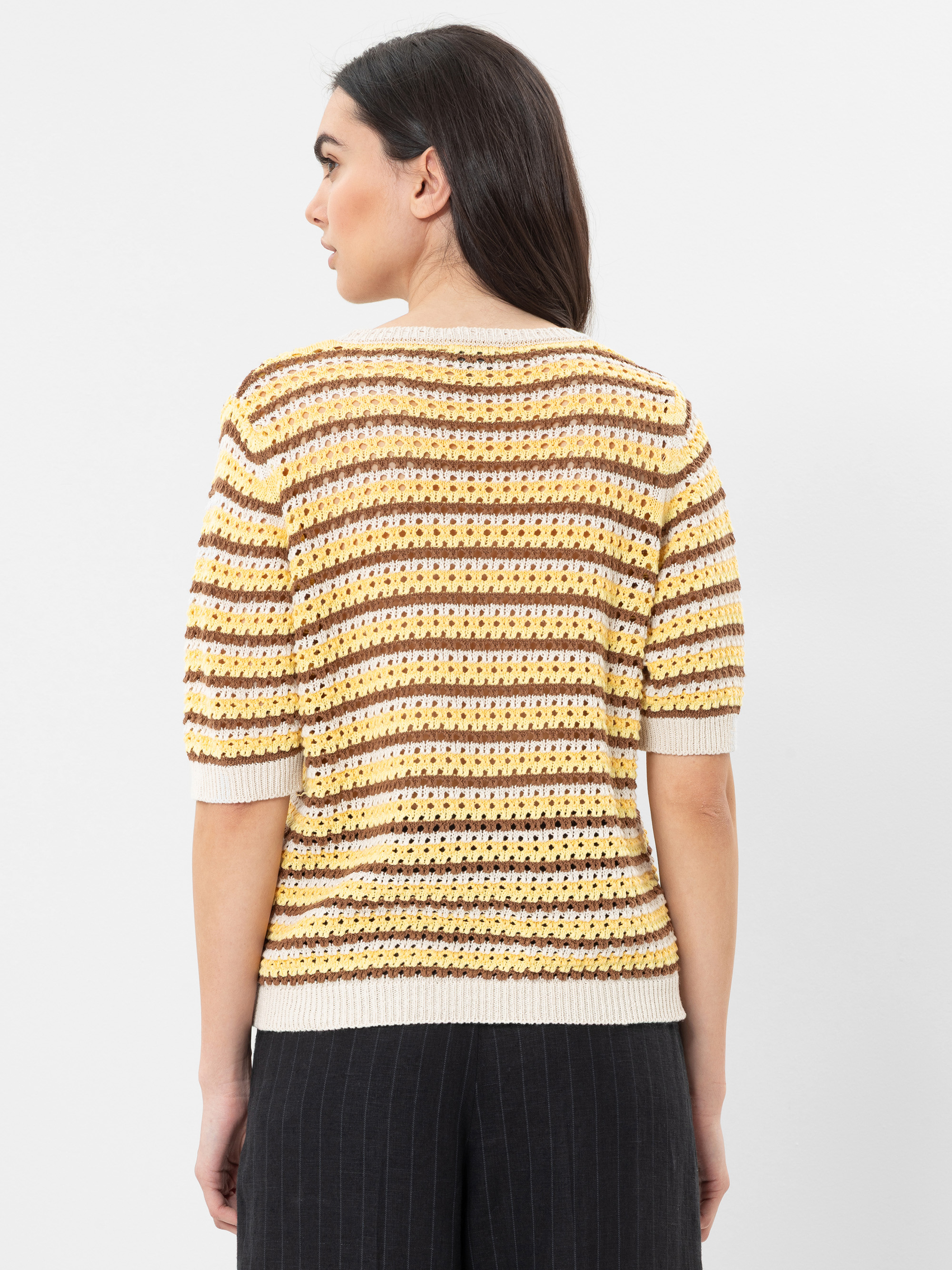 Eine Frau mit langen dunklen Haaren ist von hinten zu sehen. Sie trägt einen kurzärmeligen, gehäkelten Pullover mit horizontalen gelben, braunen und cremefarbenen Streifen, dazu eine schwarze Nadelstreifenhose.