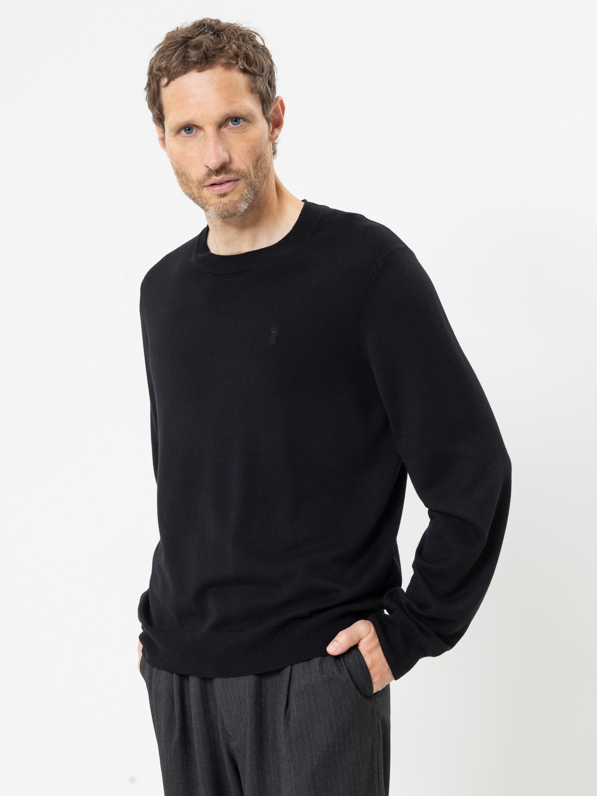 Ein Mann mit kurzen braunen Haaren und Bart trägt einen schwarzen Langarmpullover und eine graue Hose. Er steht vor einem schlichten weißen Hintergrund, die Hände in den Taschen, und blickt leicht zur Seite.