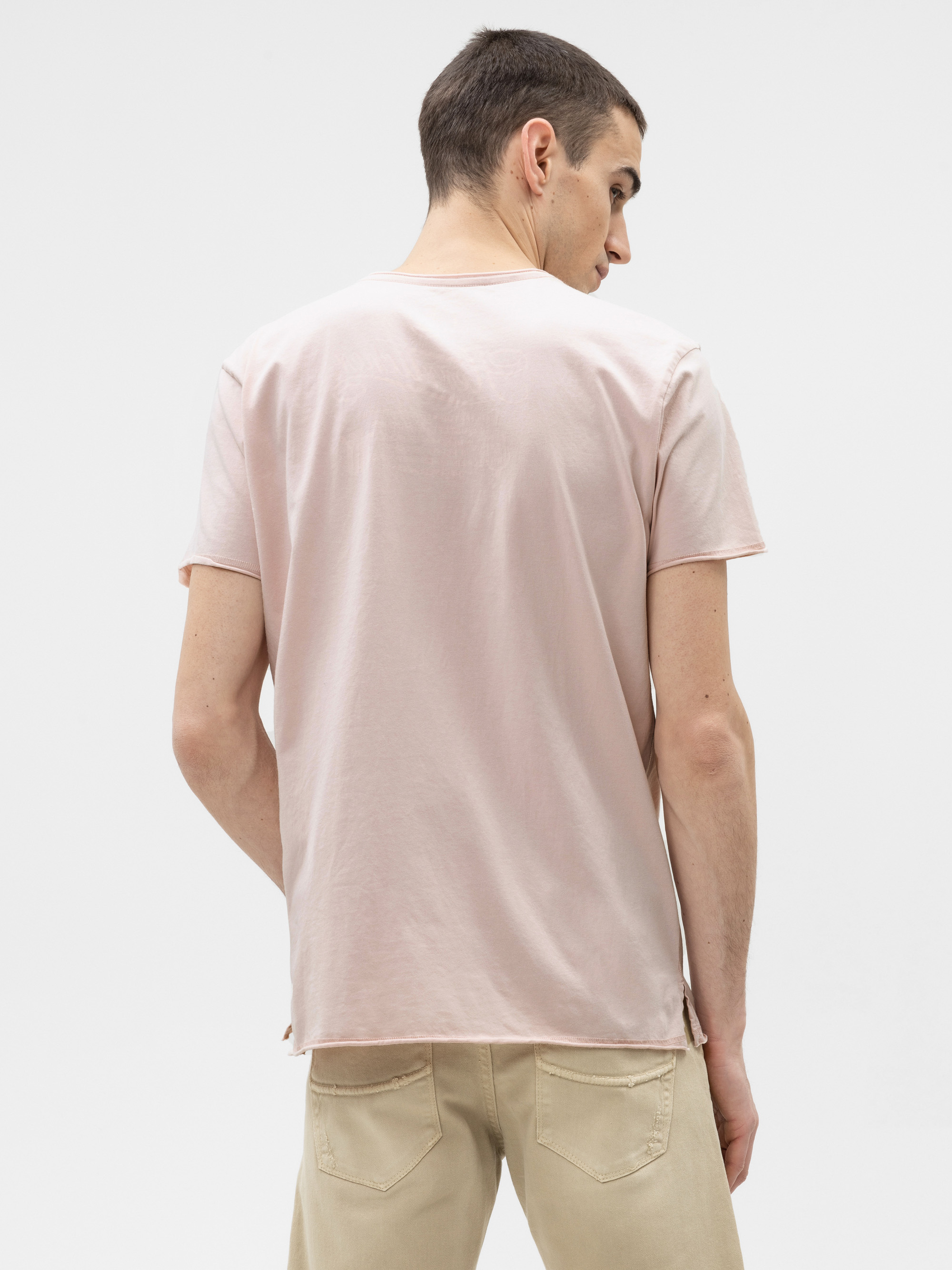 Ein Mann mit kurzen braunen Haaren wird von hinten gezeigt. Er trägt ein hellrosa kurzärmeliges T-Shirt und eine beigefarbene Hose und steht vor einem schlichten weißen Hintergrund.