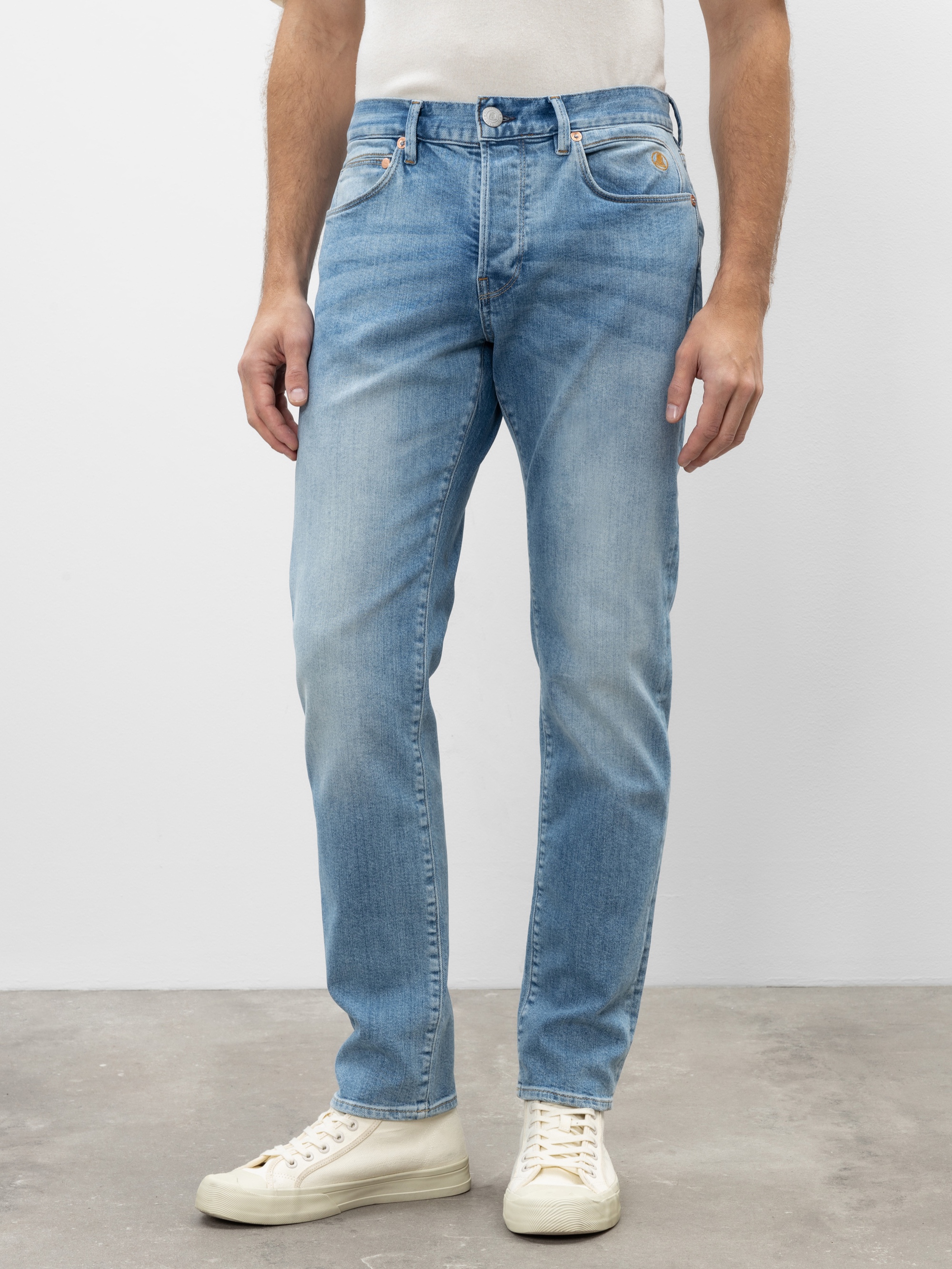 Eine Person, die hellblaue Jeans, ein weißes T-Shirt und weiße Turnschuhe trägt, steht auf einem grauen Betonboden vor einem schlichten weißen Hintergrund. Das Bild zeigt die Person von den Schultern abwärts.