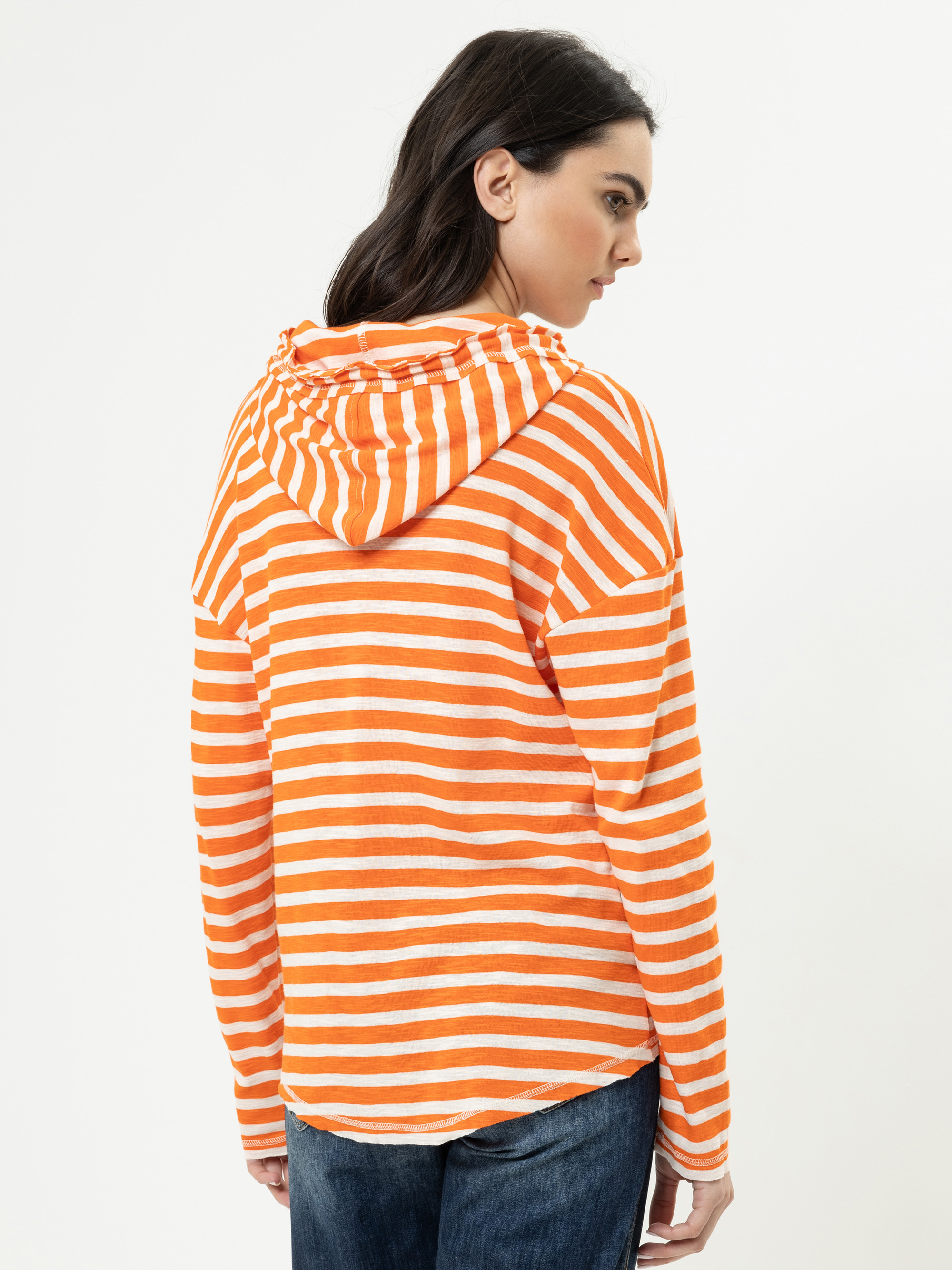 Eine Frau mit dunklem Haar trägt ein orange-weiß gestreiftes Sweatshirt mit Kapuze und blaue Jeans. Sie steht mit dem Rücken und dem Kopf leicht zur Seite gedreht vor einem schlichten weißen Hintergrund.