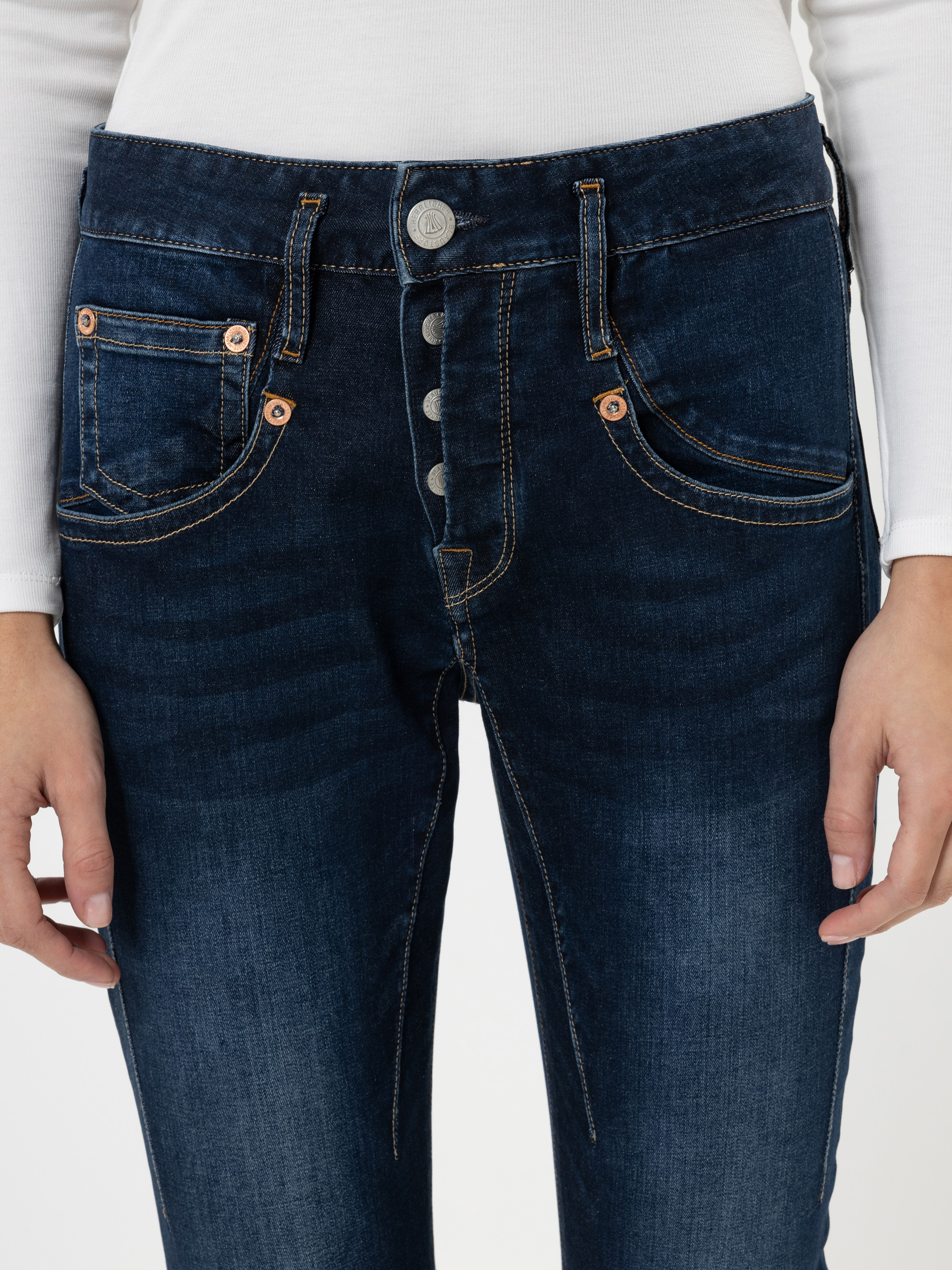 Eine Person trägt eine dunkelblaue Jeans mit Knopfleiste und sichtbaren hellbraunen Nähten, dazu ein weißes langärmeliges Oberteil. Die Hände der Person ruhen an der Seite.