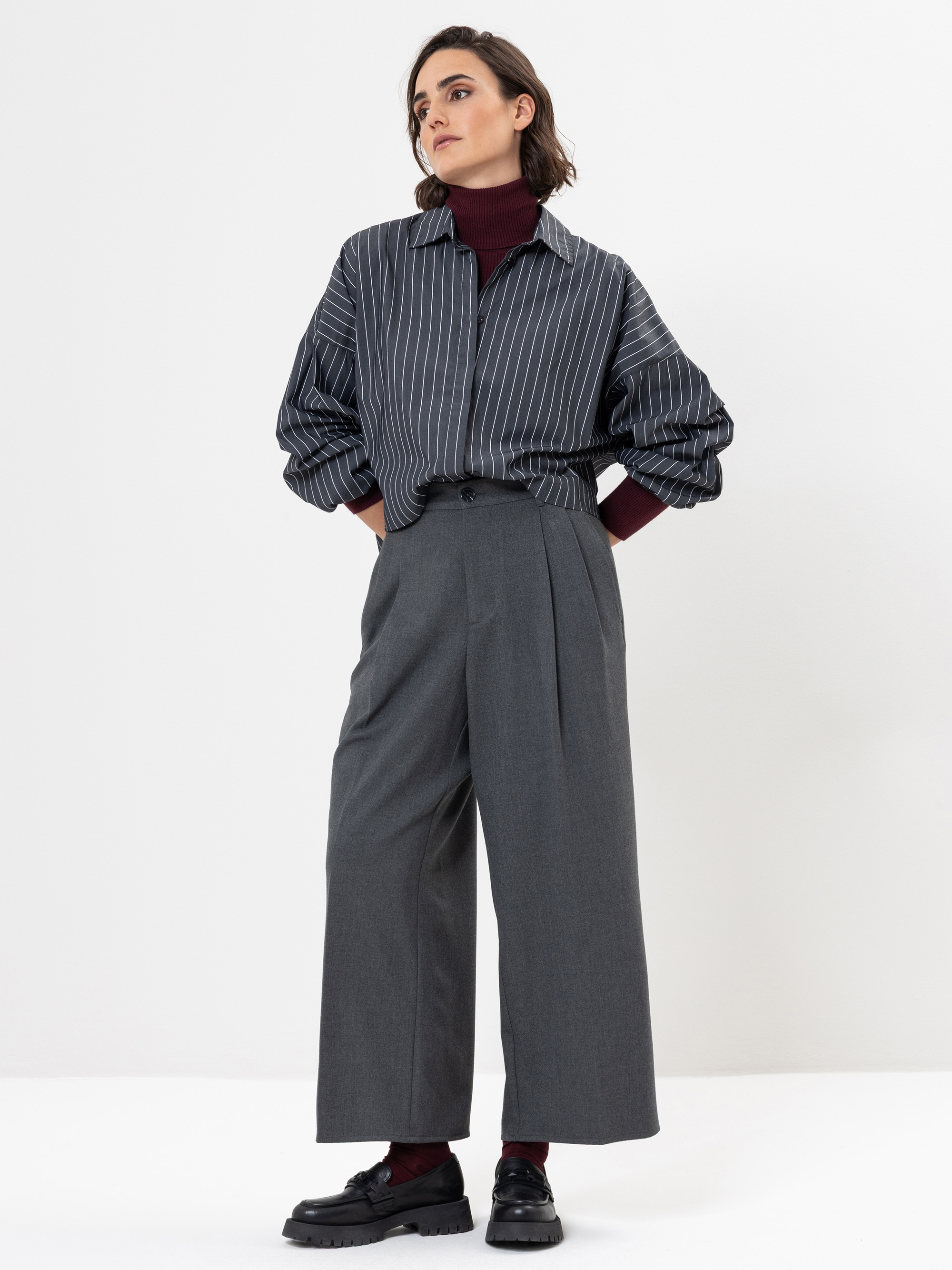 Eine Frau steht vor einem weißen Hintergrund und trägt ein graues Nadelstreifenhemd, einen dunkelroten Rollkragenpullover, eine graue Hose mit weitem Bein und schwarze Schuhe. Sie hat kurzes braunes Haar und schaut mit den Händen in den Taschen zur Sei