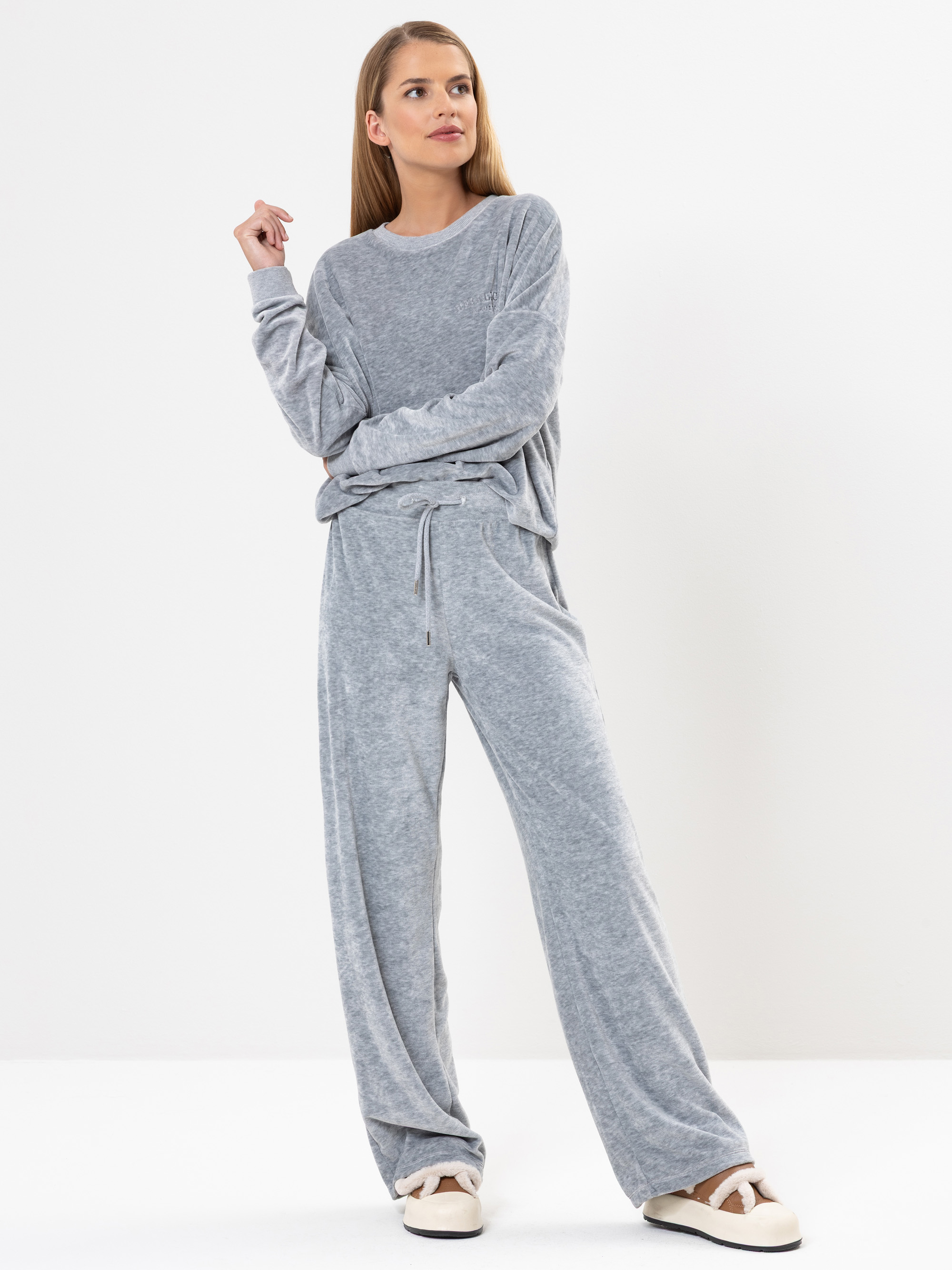 Eine Frau steht vor einem weißen Hintergrund und trägt ein locker sitzendes, hellgraues Loungewear-Set mit langen Ärmeln und einer Hose mit Kordelzug, gepaart mit weißen Hausschuhen. Sie blickt mit entspanntem Blick zur Seite.
