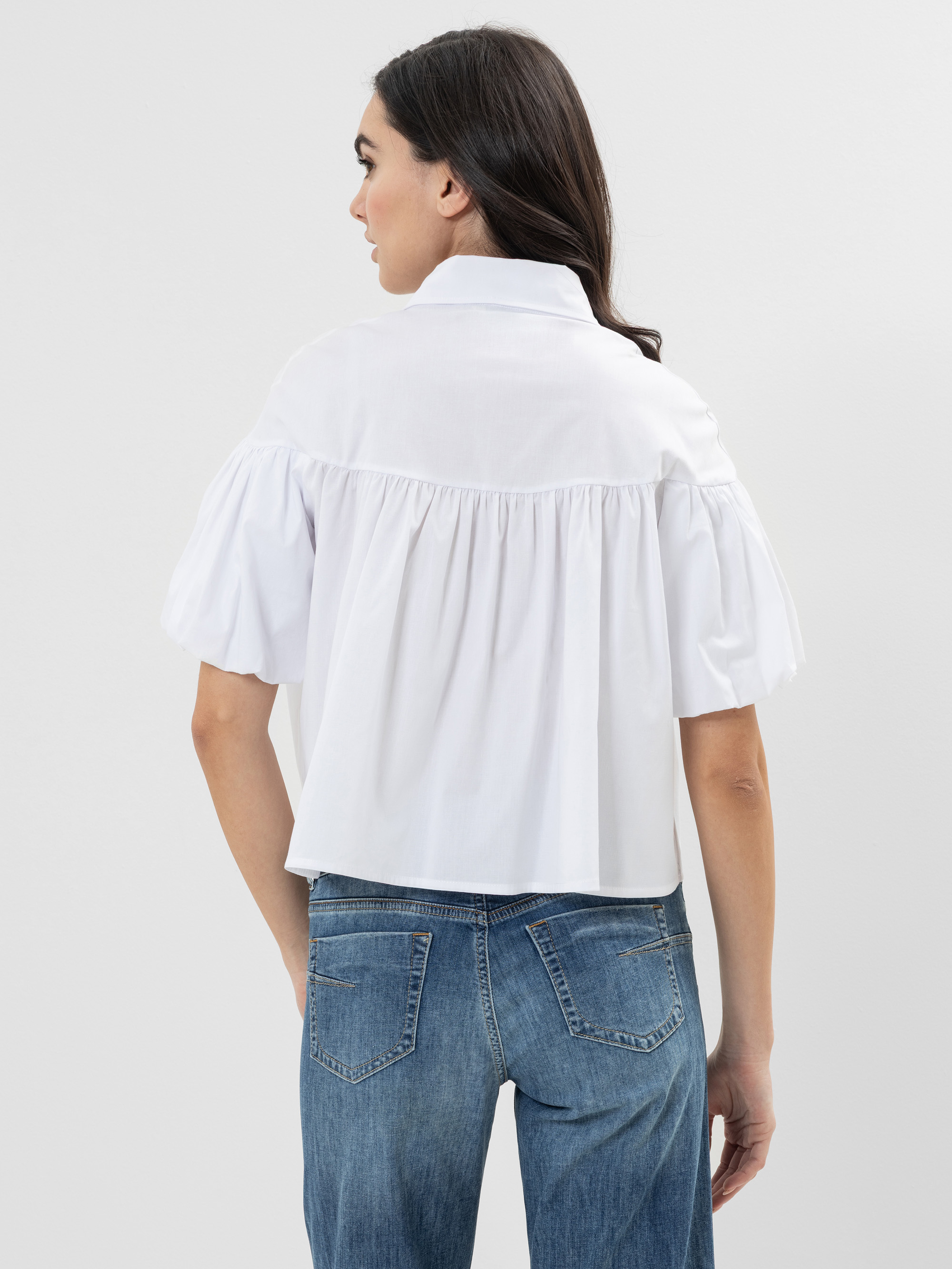 Frau mit Blickrichtung nach hinten, trägt eine weiße kurzärmelige Bluse mit gerafftem Stoff und blaue Jeans. Ihr dunkles Haar ist offen, und der Hintergrund ist einfarbig hellgrau.