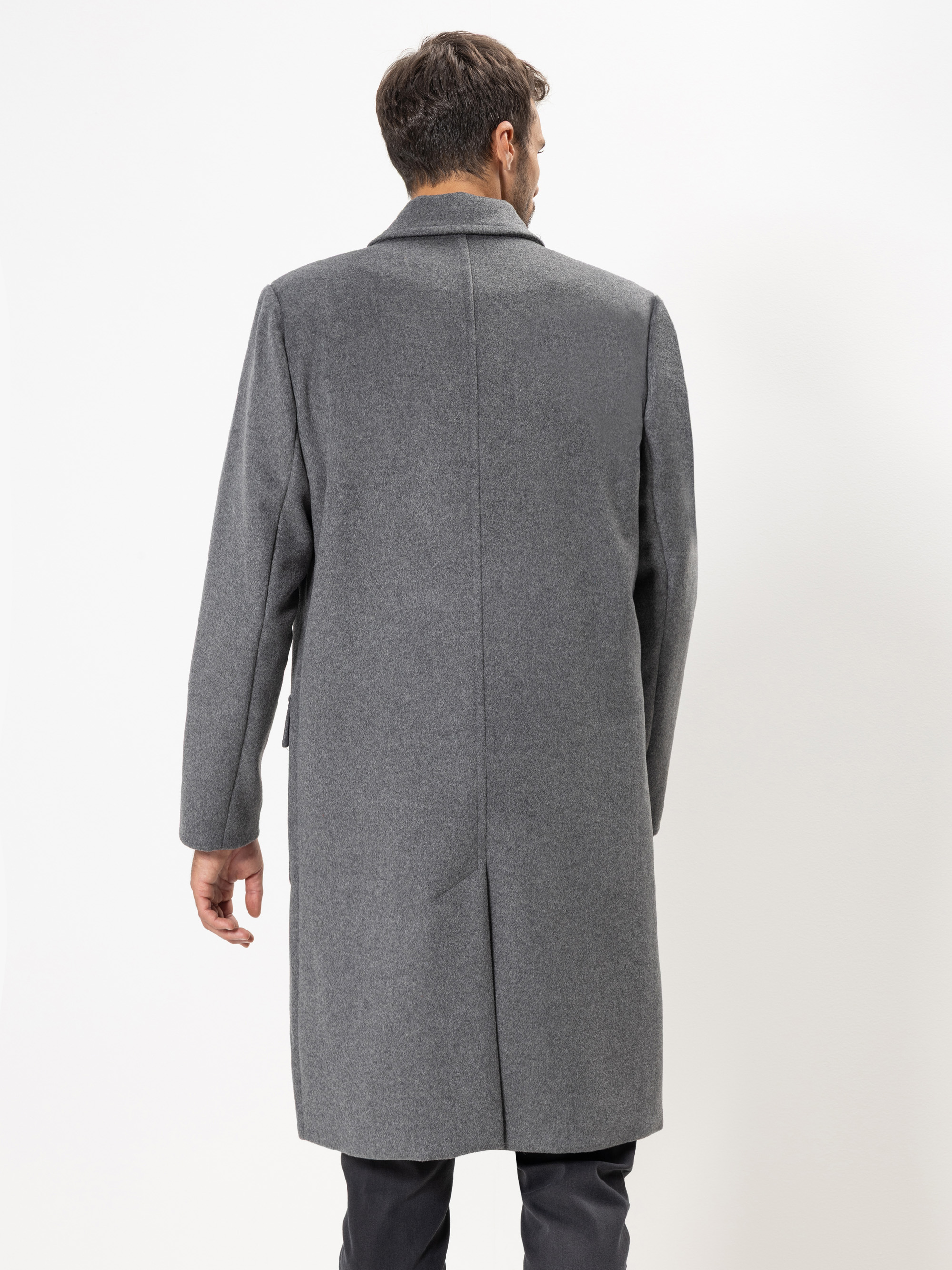 Ein Mann in einem langen grauen Mantel steht mit dem Gesicht von der Kamera weg vor einem schlichten weißen Hintergrund.