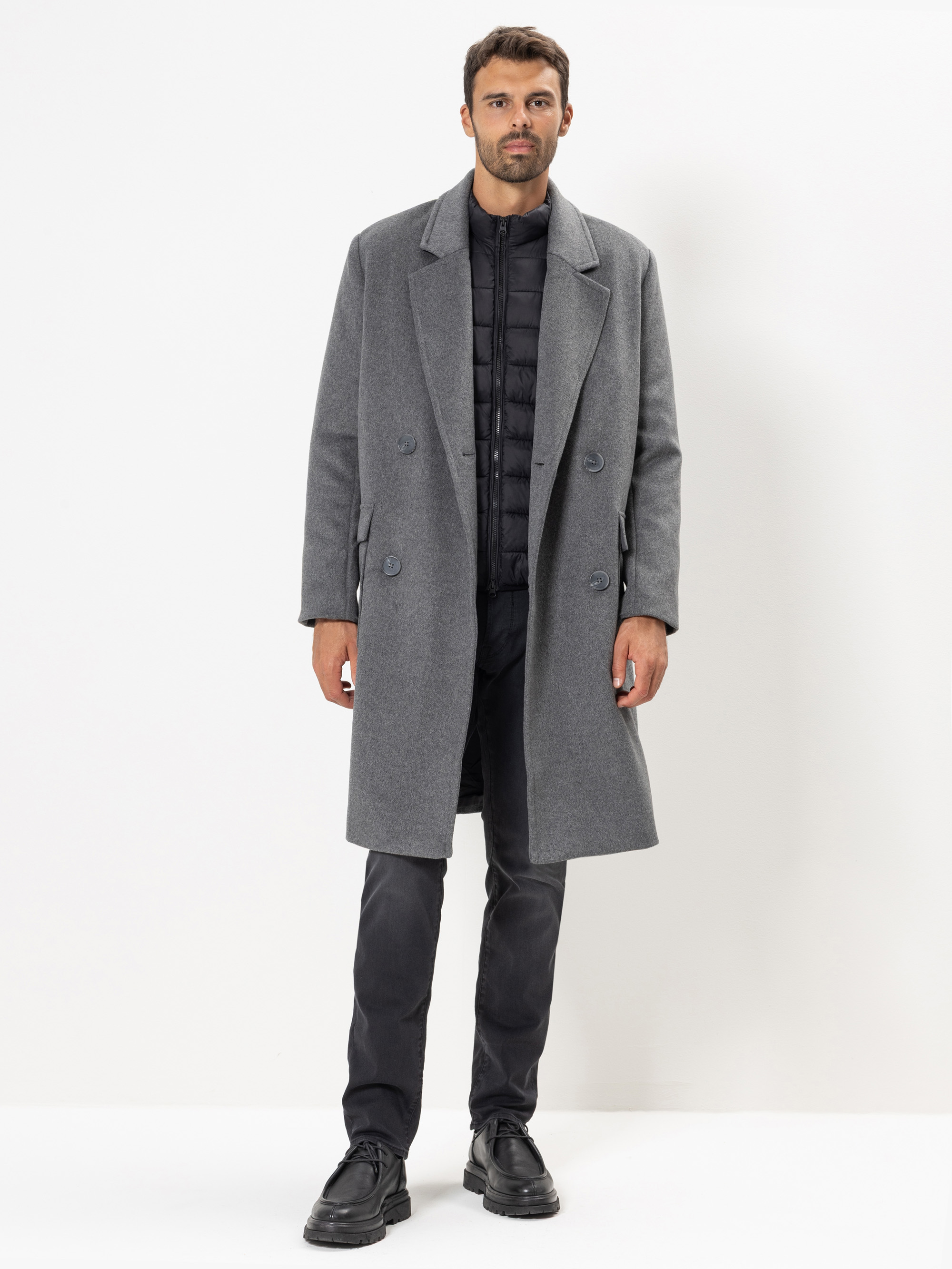Ein Mann steht mit dem Gesicht nach vorne vor einem weißen Hintergrund. Er trägt einen langen grauen Mantel über einer schwarzen Steppjacke, schwarze Hosen und schwarze Anzugschuhe.