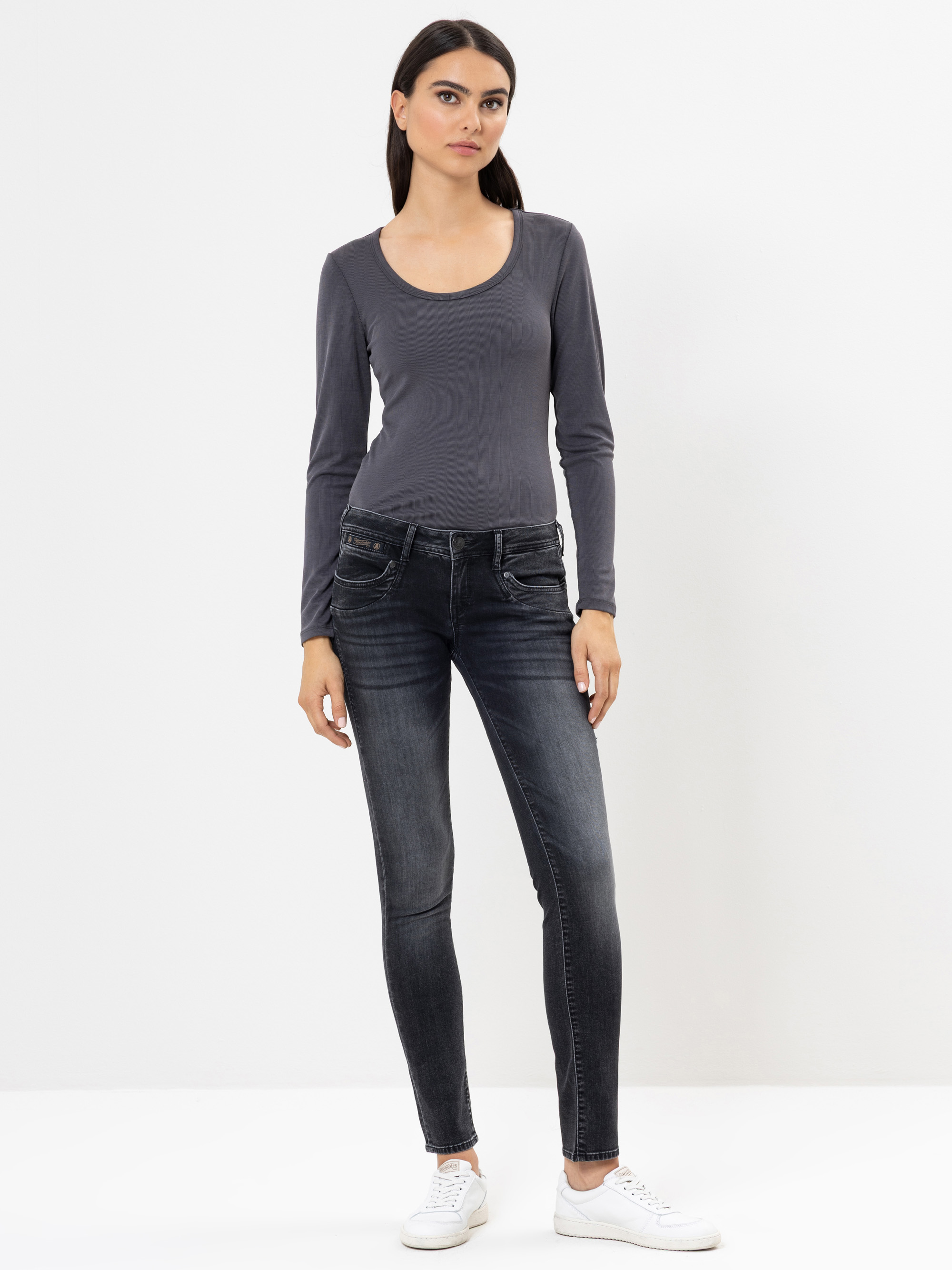 Eine Frau steht vor einem schlichten weißen Hintergrund und trägt ein langärmeliges graues Oberteil, dunkle Röhrenjeans und weiße Turnschuhe. Sie hat glattes dunkles Haar und einen neutralen Gesichtsausdruck.